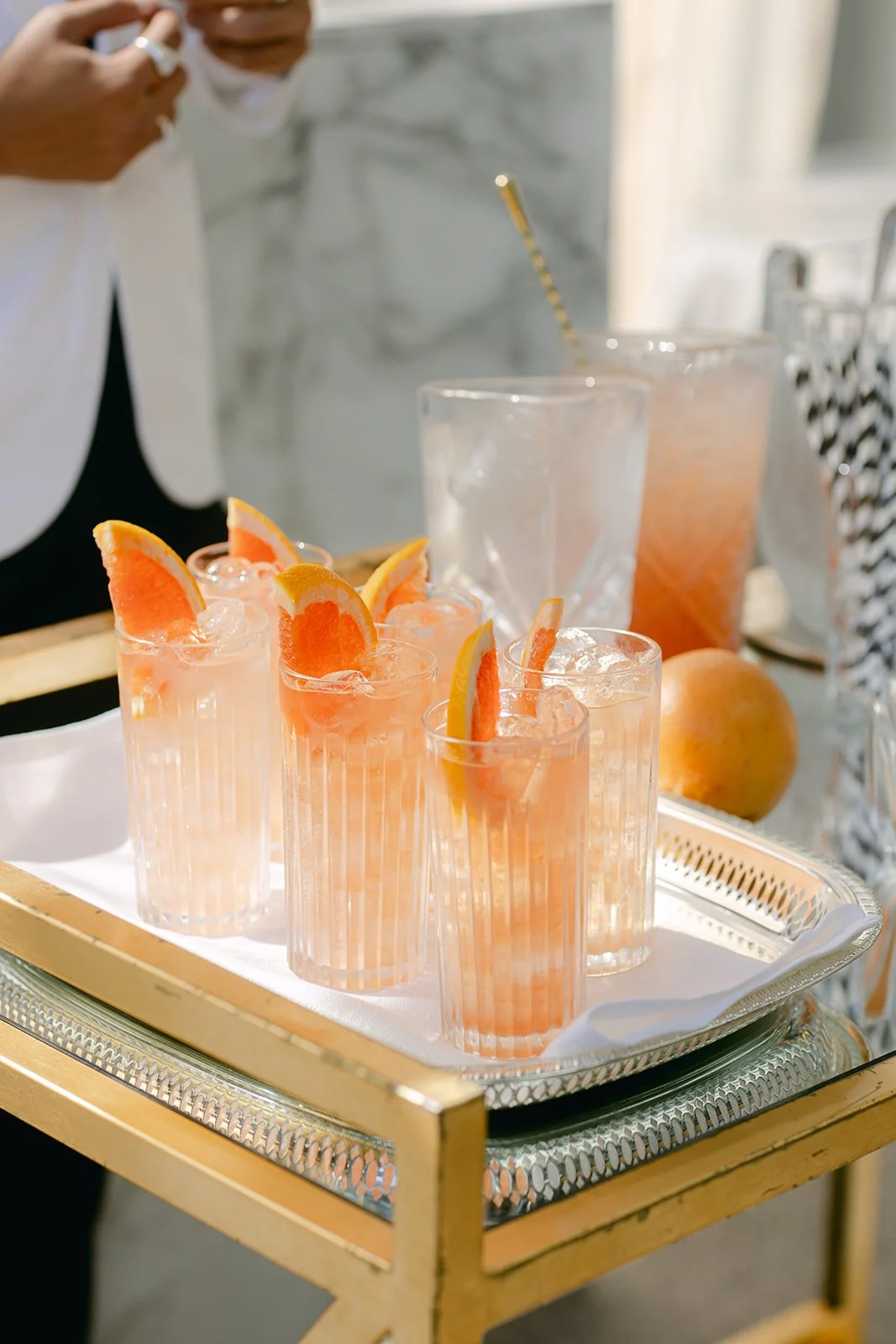 A silver tray with six glasses of grapefruit cocktails garnished with grapefruit and lemon wedges. The tray is on a small, gold-colored table. Two glasses of water and some oranges are in the background.