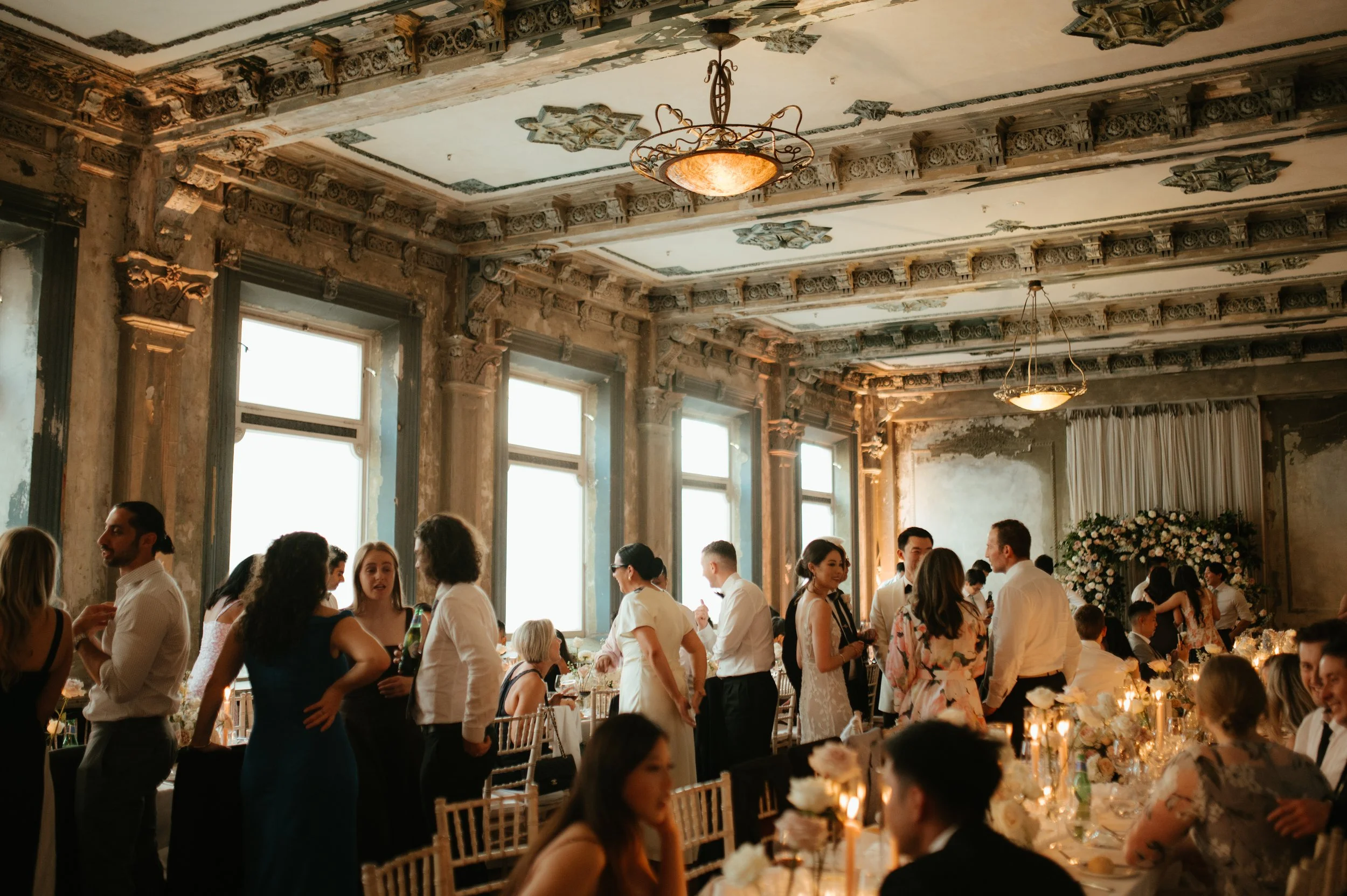 Guests mingling at a wedding reception in a vintage-style decorated hall with high ceilings, ornate moldings, and large windows.