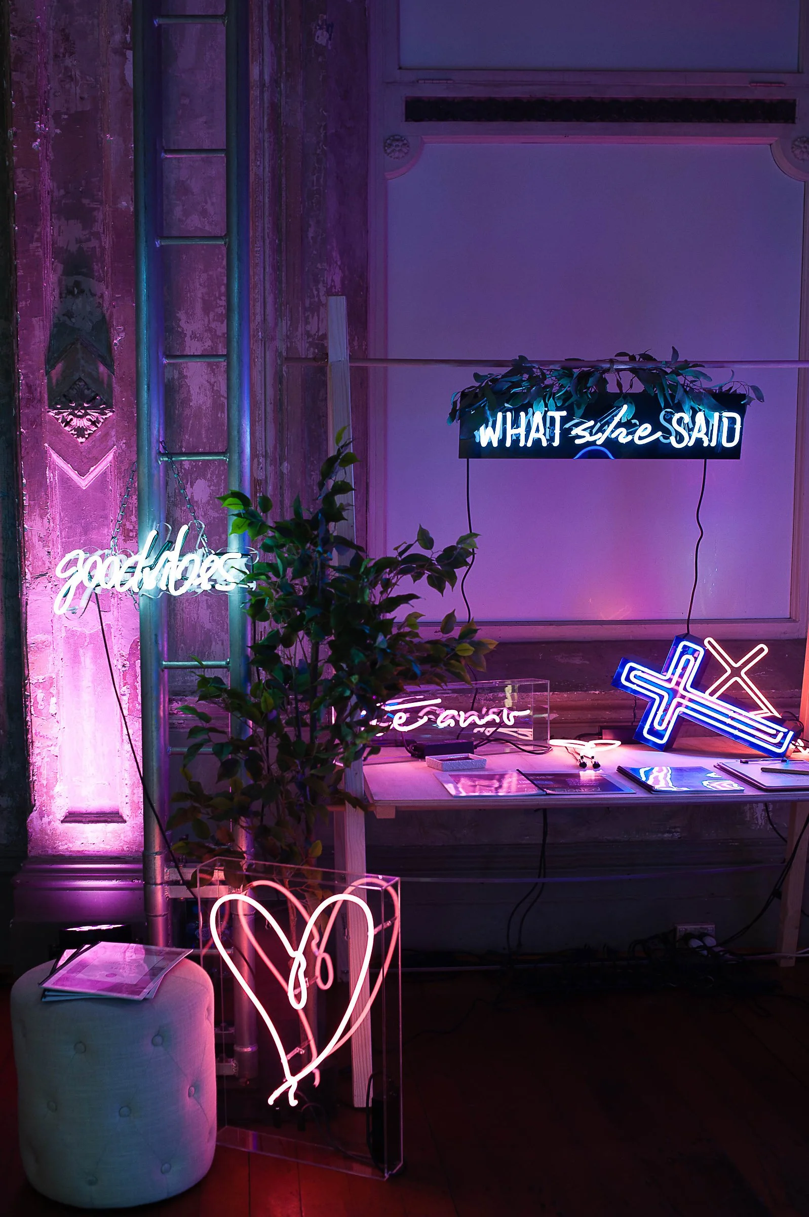 Neon signs reading 'What she said,' 'good vibes,' and a heart with an arrow, along with a cross and other neon decor in a dimly lit room with pink and purple lighting, a plant, books, and a small white ottoman.