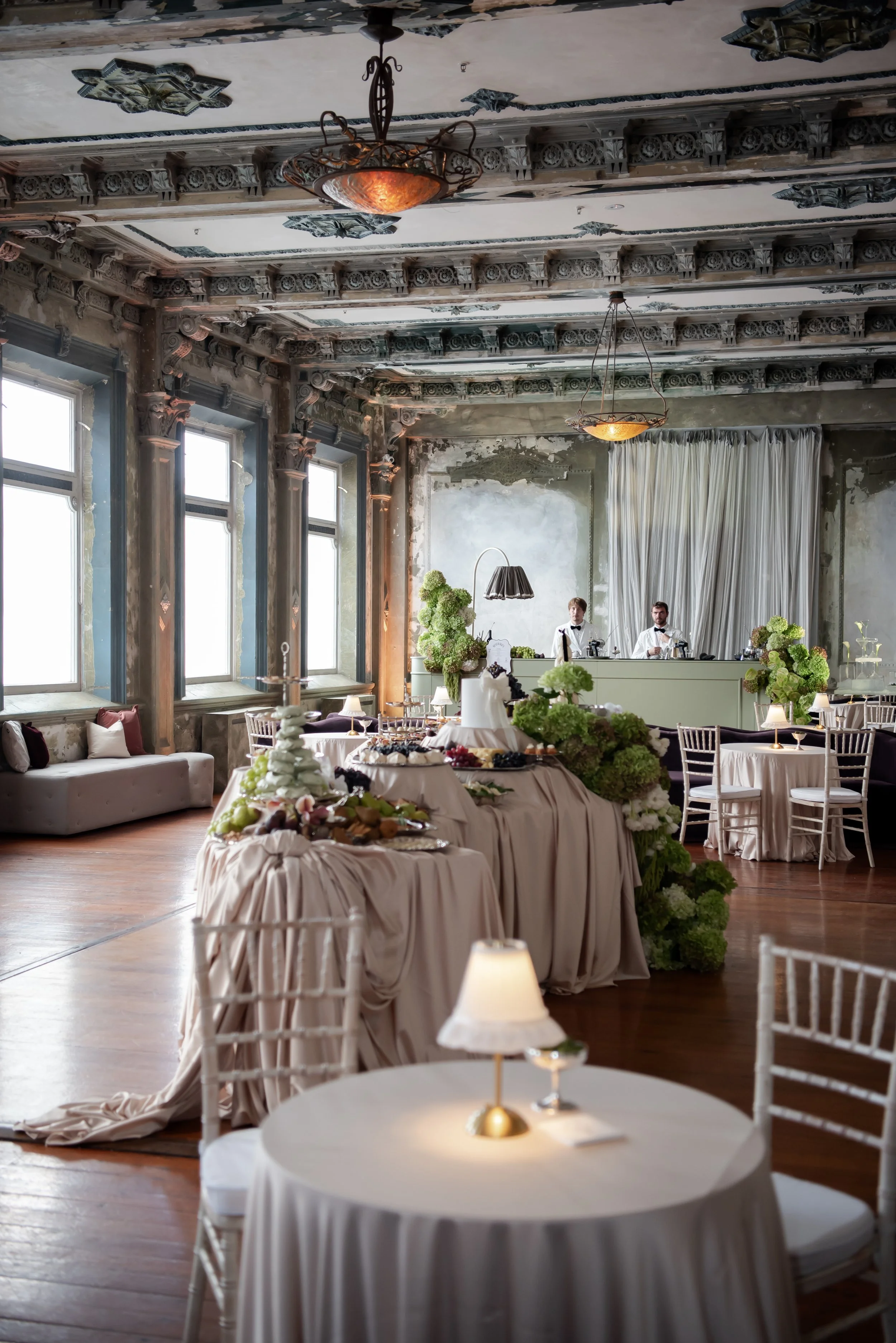 Elegant event space with ornate ceiling, large windows, draped curtains, and decorated tables with floral arrangements and desserts.