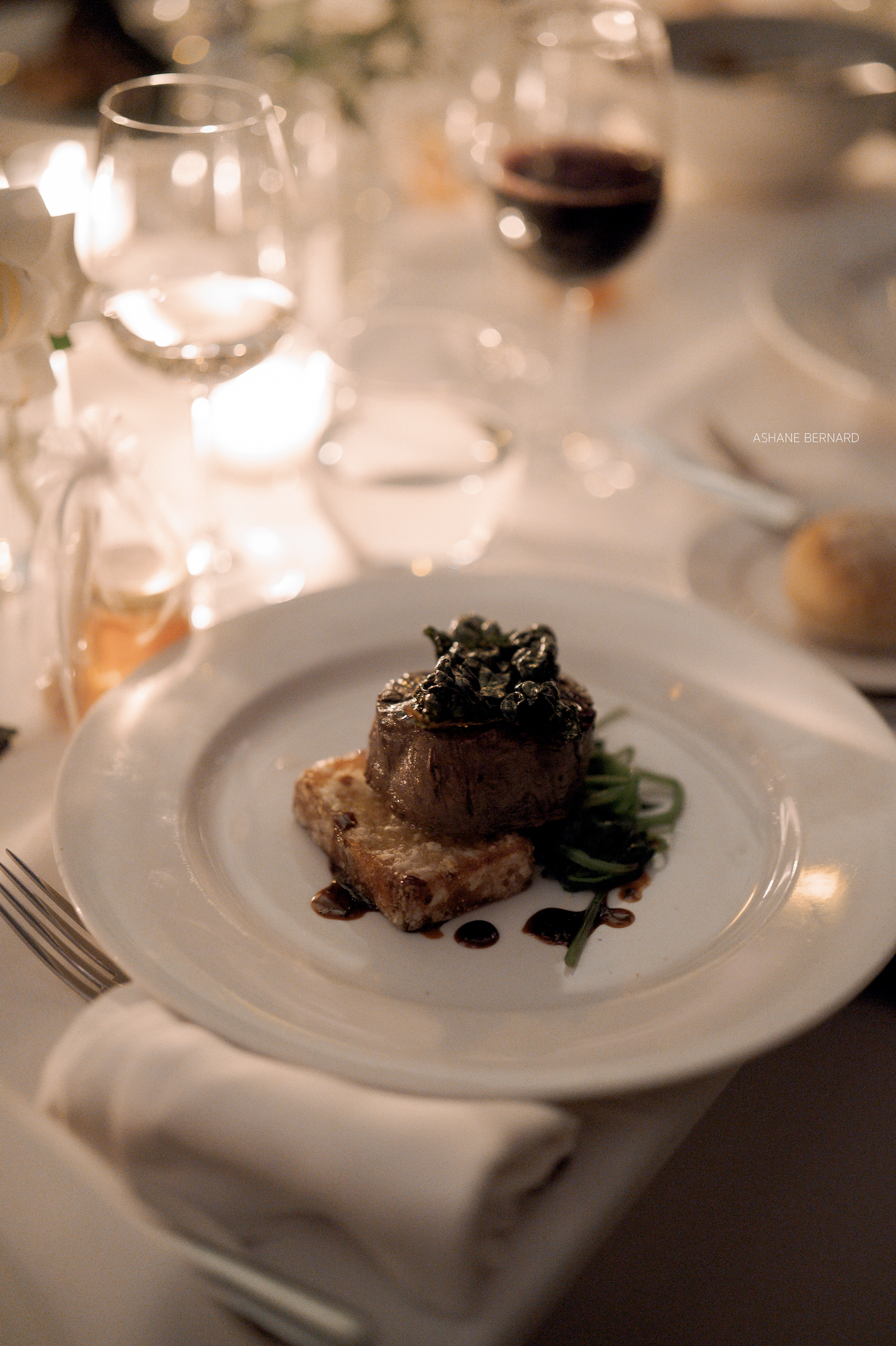 A plated gourmet dish featuring a piece of steak topped with black caviar, served on a white plate with a side of greens, set on a table with wine glasses and candlelight.