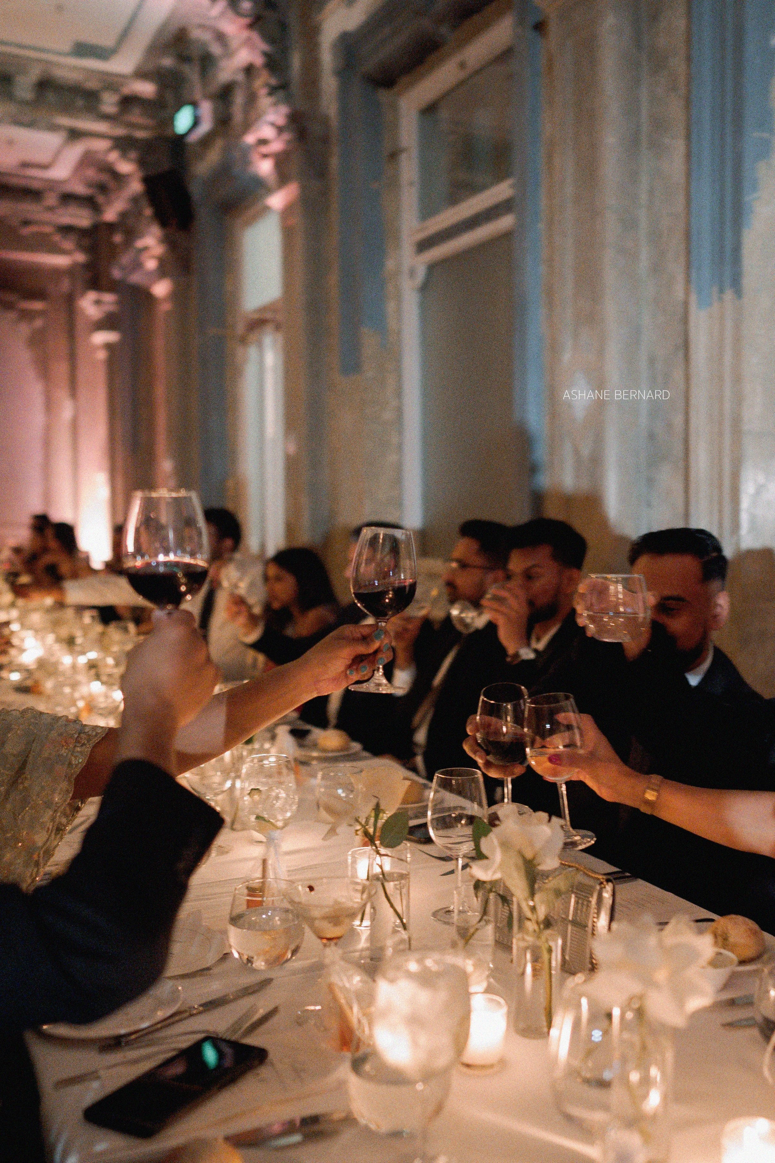 People at a formal dinner raising glasses of wine in a toast, sitting at a decorated table with candles and flowers in an elegant setting.
