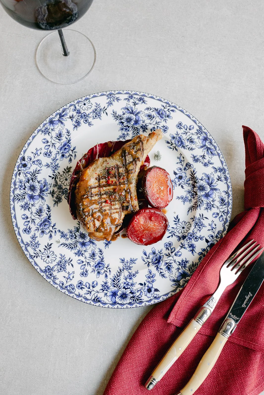A plate with grilled meat, glazed carrots, and red beets, accompanied by a glass of red wine, set on a table with a red napkin and silverware.