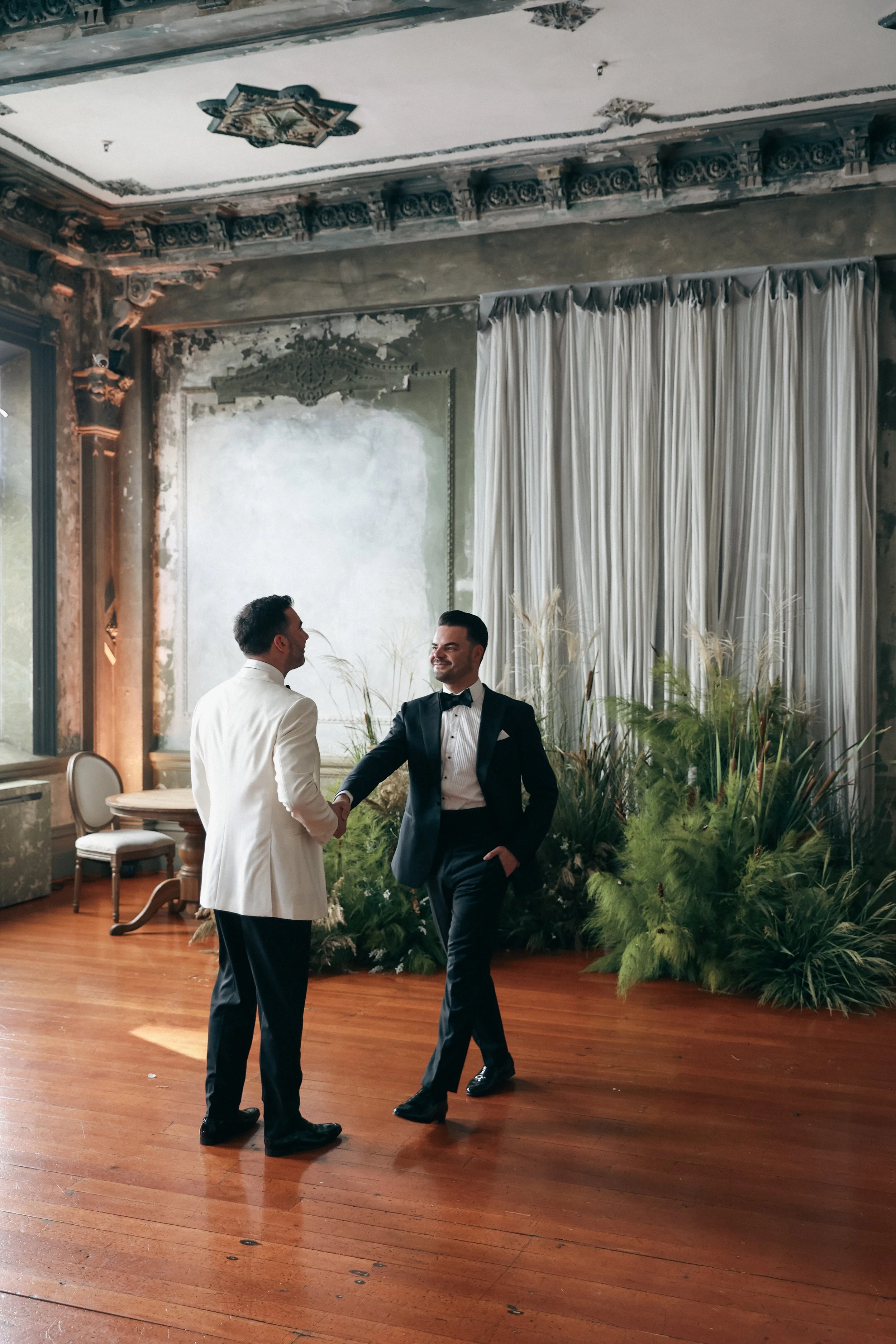 Two grooms dressed in tuxedos shaking hands in an elegantly decorated room with wooden floors, ornate ceiling details, the George Ballroom