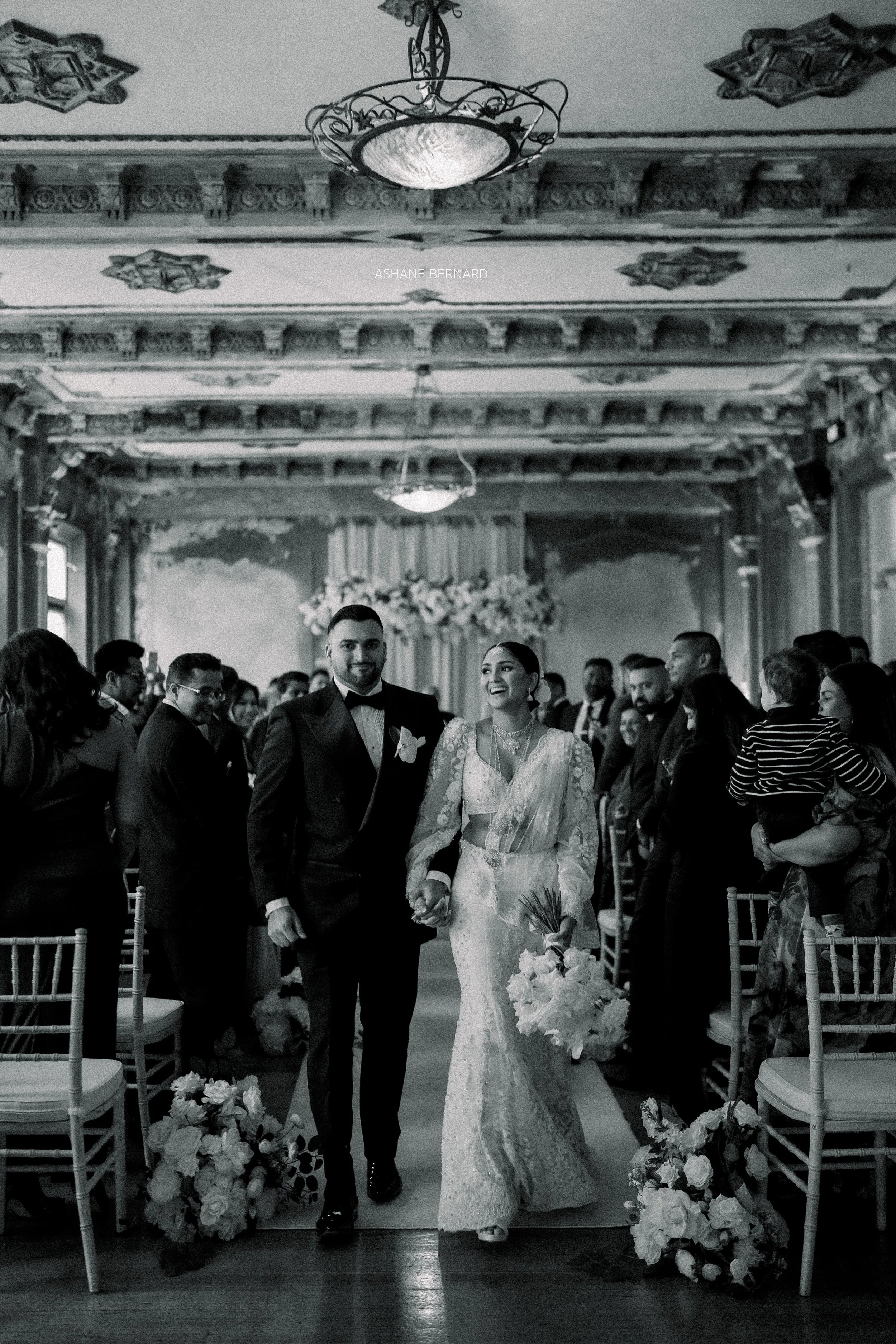 A bride and groom walking down the aisle at their wedding ceremony in an elegant indoor venue, surrounded by guests and floral arrangements.