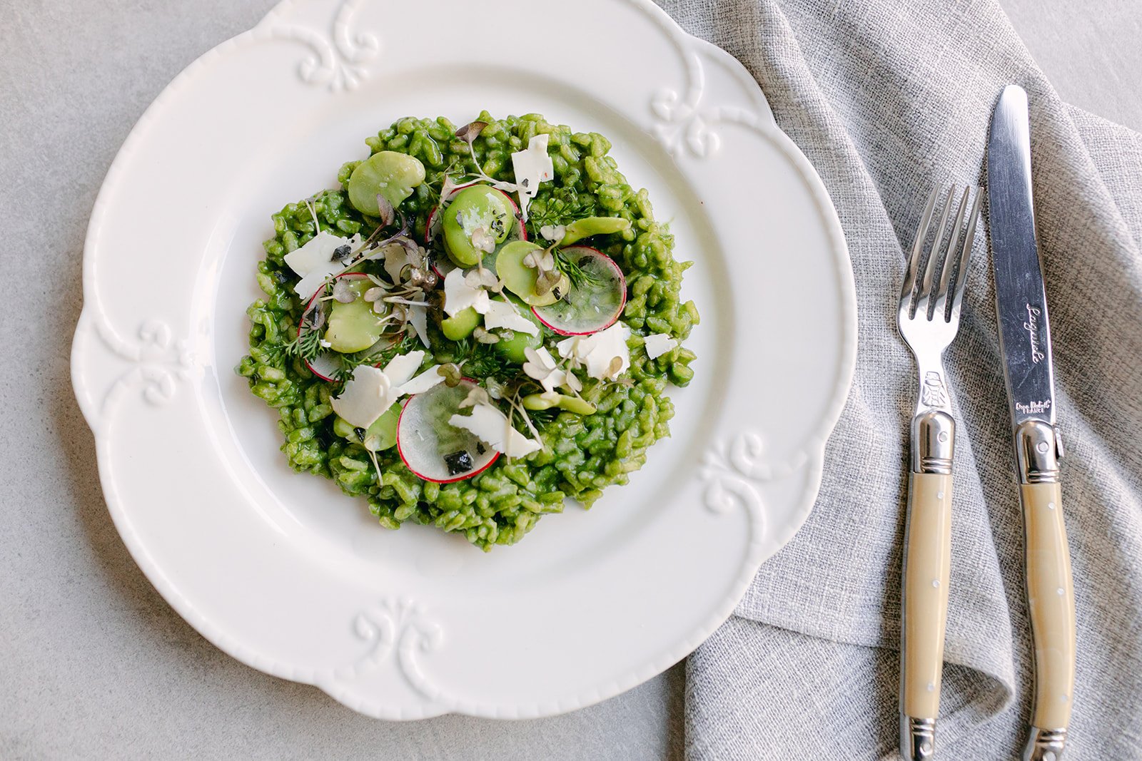 A plate of green risotto garnished with radish slices, microgreens, and shaved cheese, placed on a gray tablecloth with a fork and knife beside it.