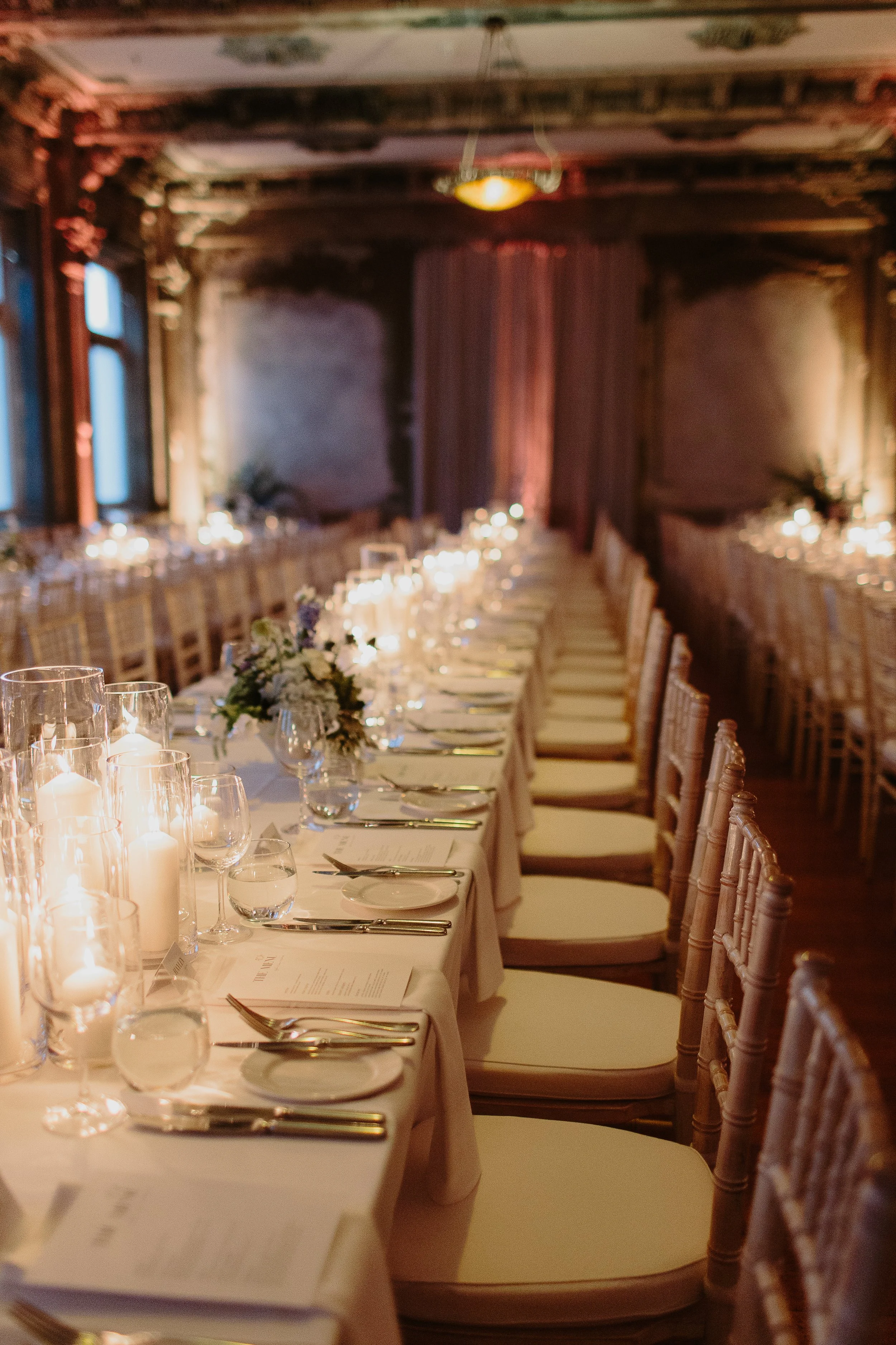 Elegant banquet table set with white linens, multiple candles, floral centerpieces, glassware, silverware, and printed menus in a warmly lit, vintage-style room with wooden beams and large windows.