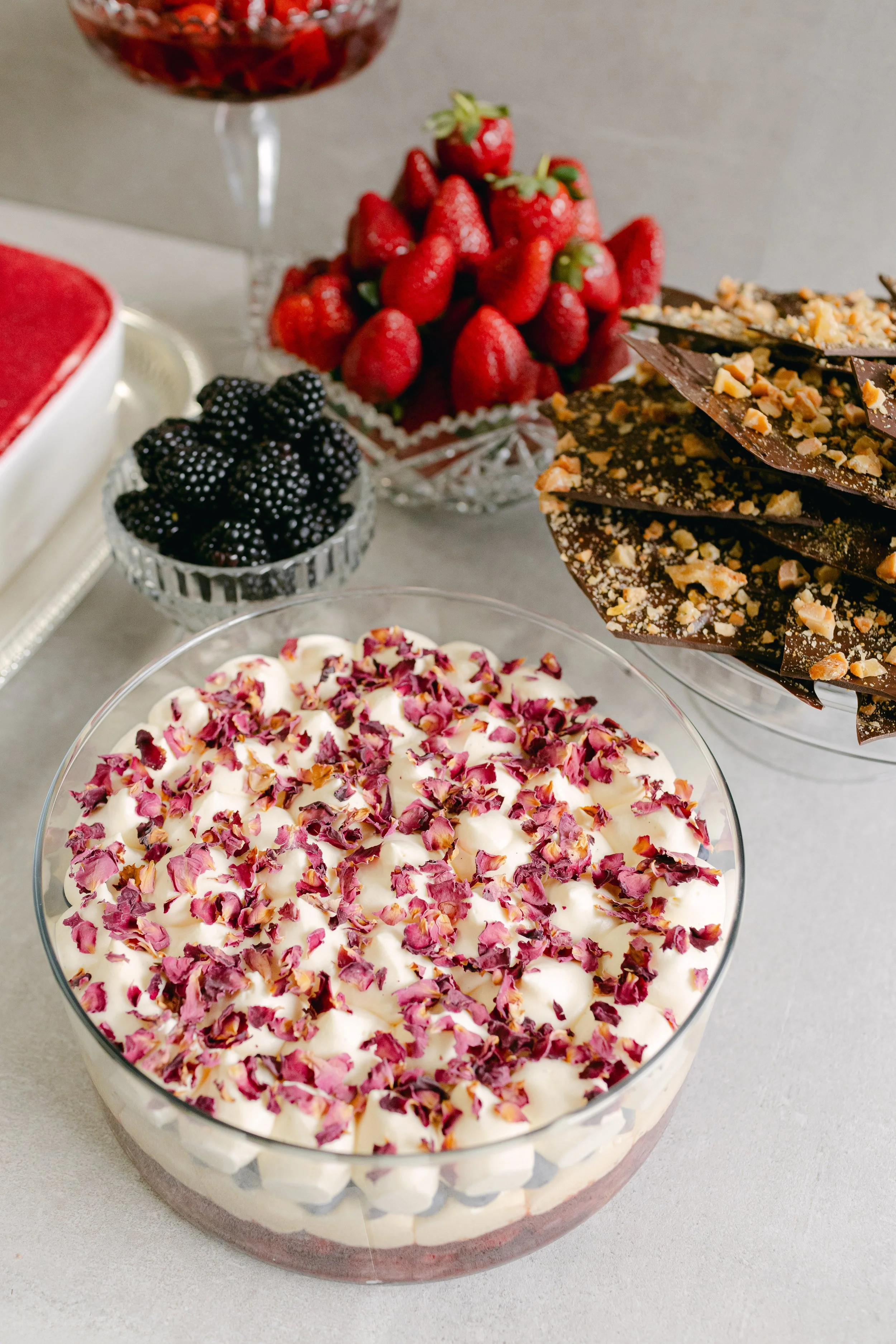 Assorted desserts with fresh berries, including a layered berry trifle topped with rose petals, a bowl of blackberries, a bowl of strawberries, and chocolate with chopped nuts.