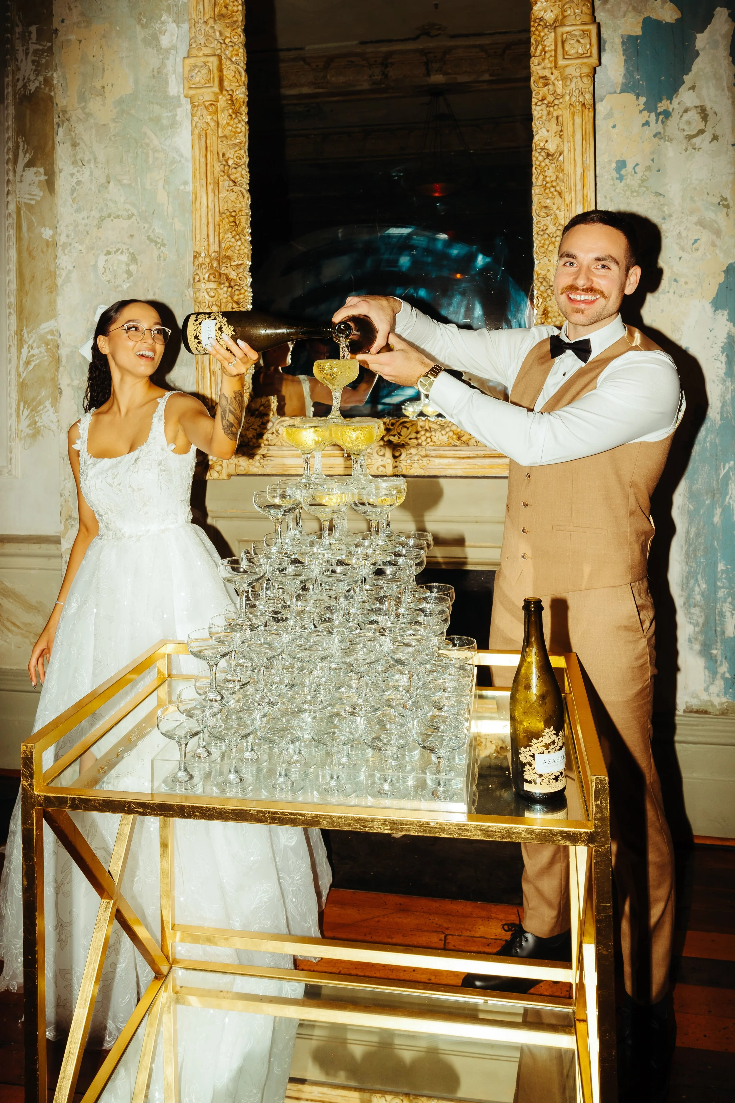 A bride and groom pour champagne into a pyramid of glasses at their wedding reception.