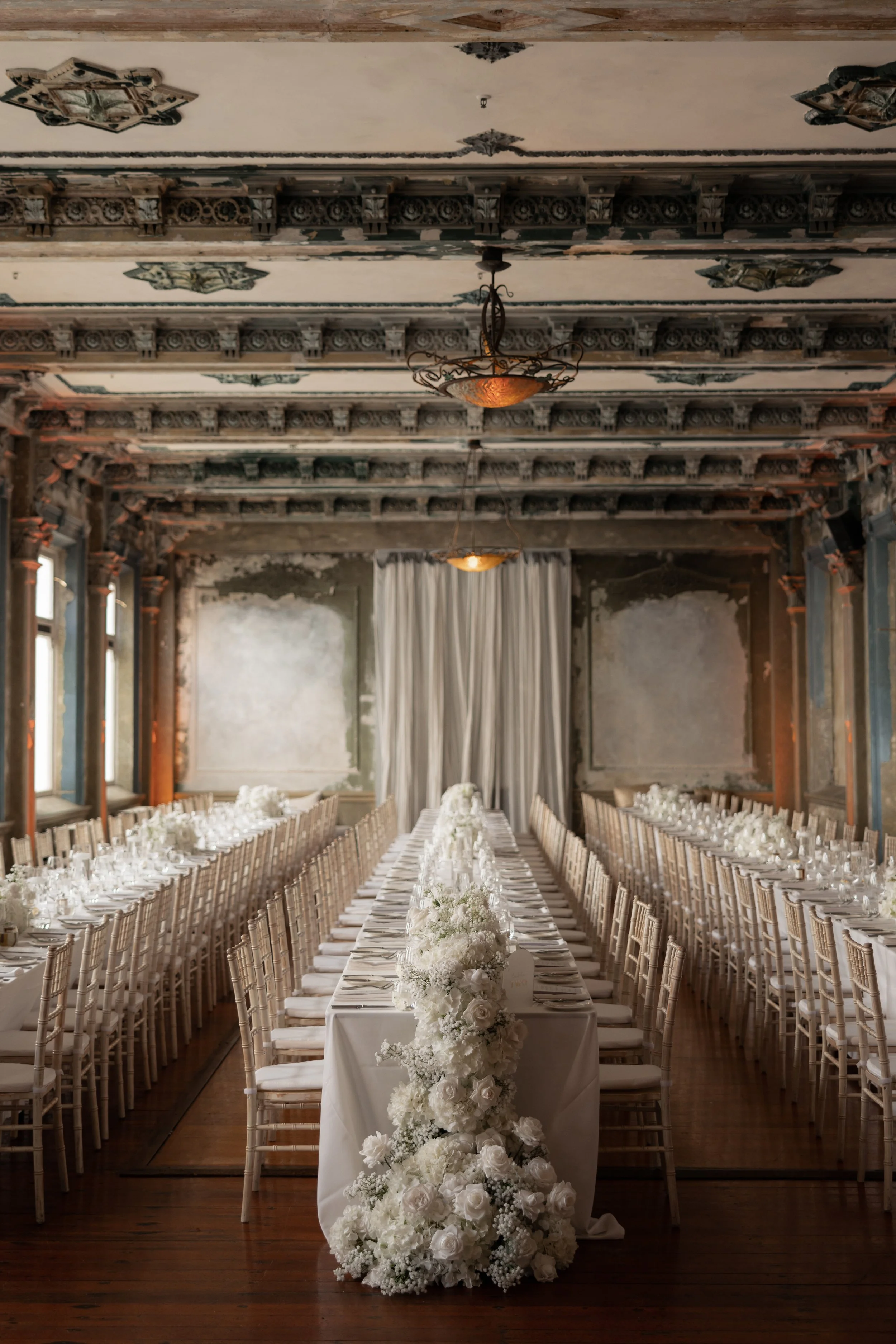 Elegant wedding reception setup with long white tables, white chairs, and floral centerpieces, in a vintage room with ornate ceiling and large windows.