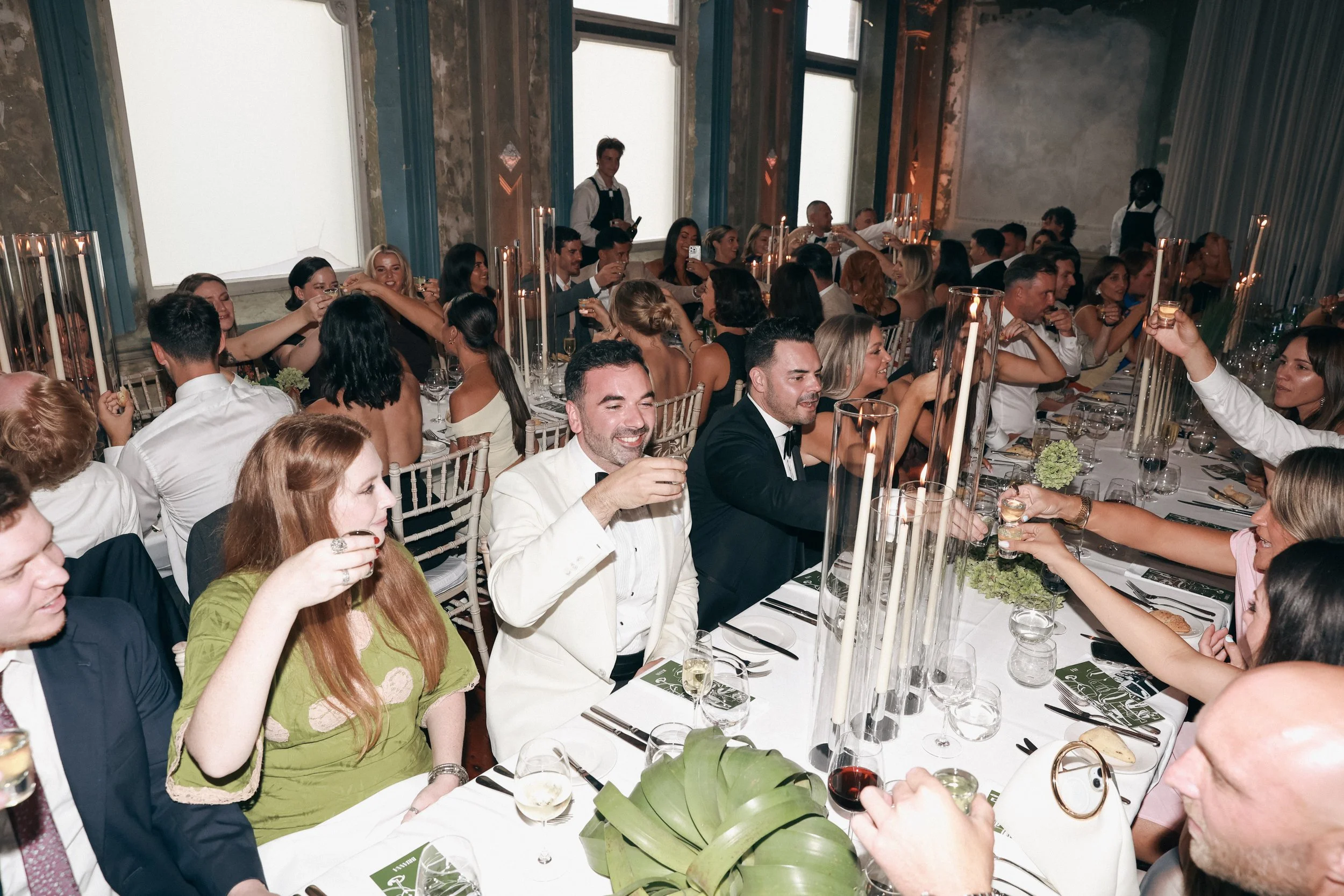 People celebrating at a formal dinner event with a long table, floral centerpieces, candlelit ambiance, and many guests raising glasses in a toast.