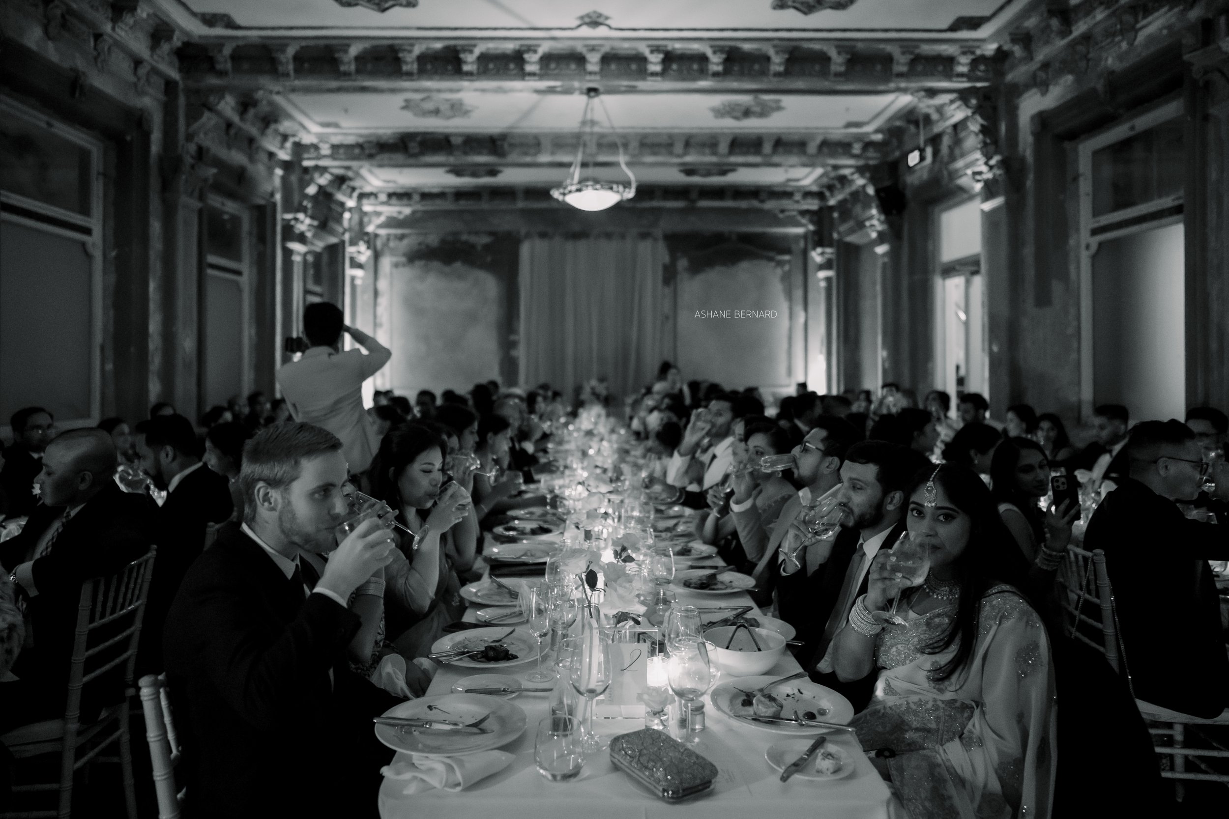People at a formal dinner party in an elegant, ornate hall, sitting at a long table with food and drinks, some are taking photos, black and white image.