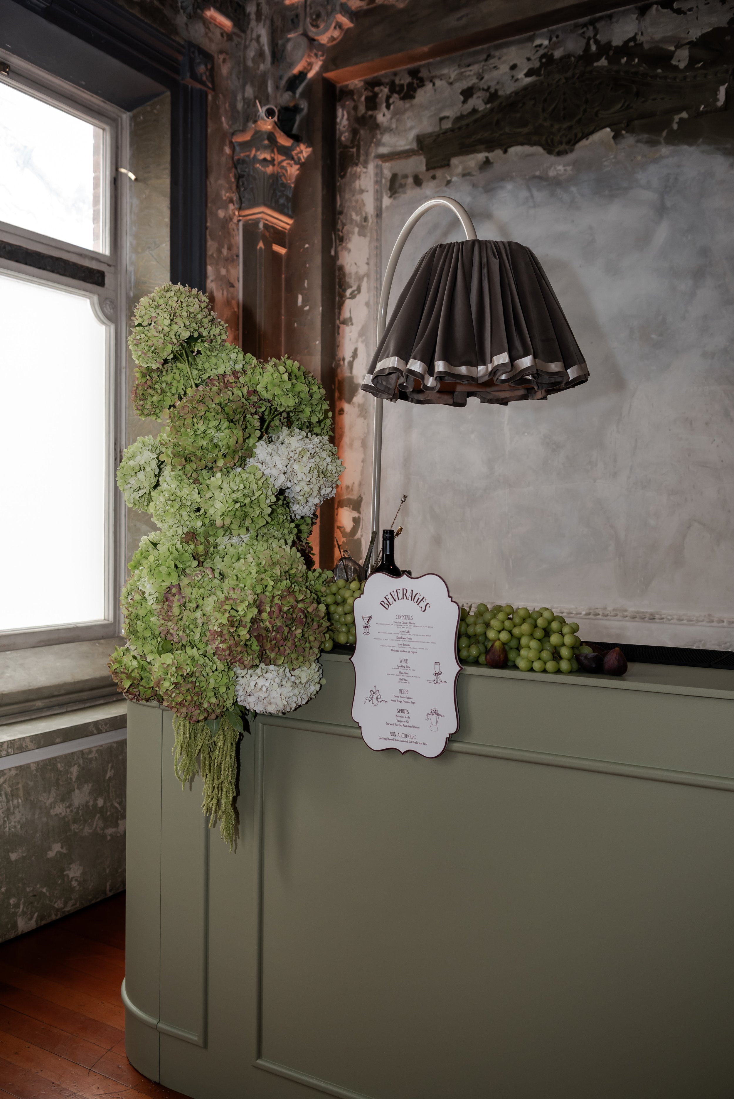 A vintage bar with a gray curved front and a large floral arrangement of green and white hydrangeas, a black lampshade, and a beverage menu, set against a distressed wall with a window to the left.