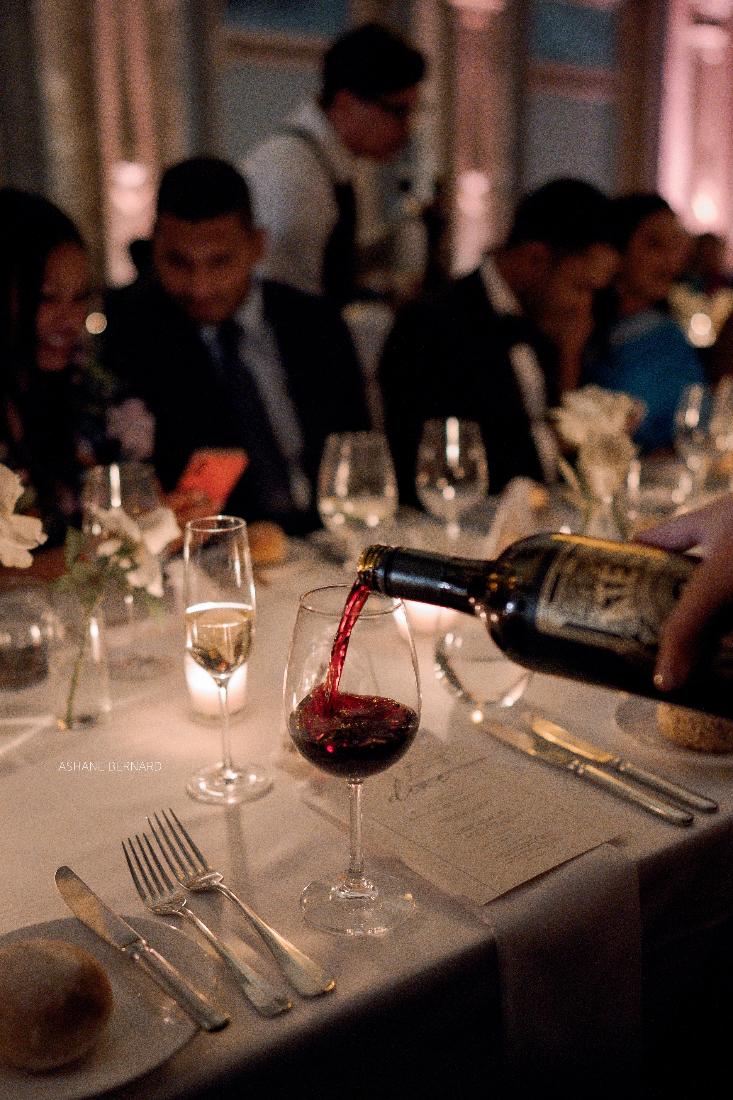 A person is pouring red wine into a wine glass at a formal dinner event. The table is set with silverware, glassware, and a menu, with people dressed in formal attire seated in the background.