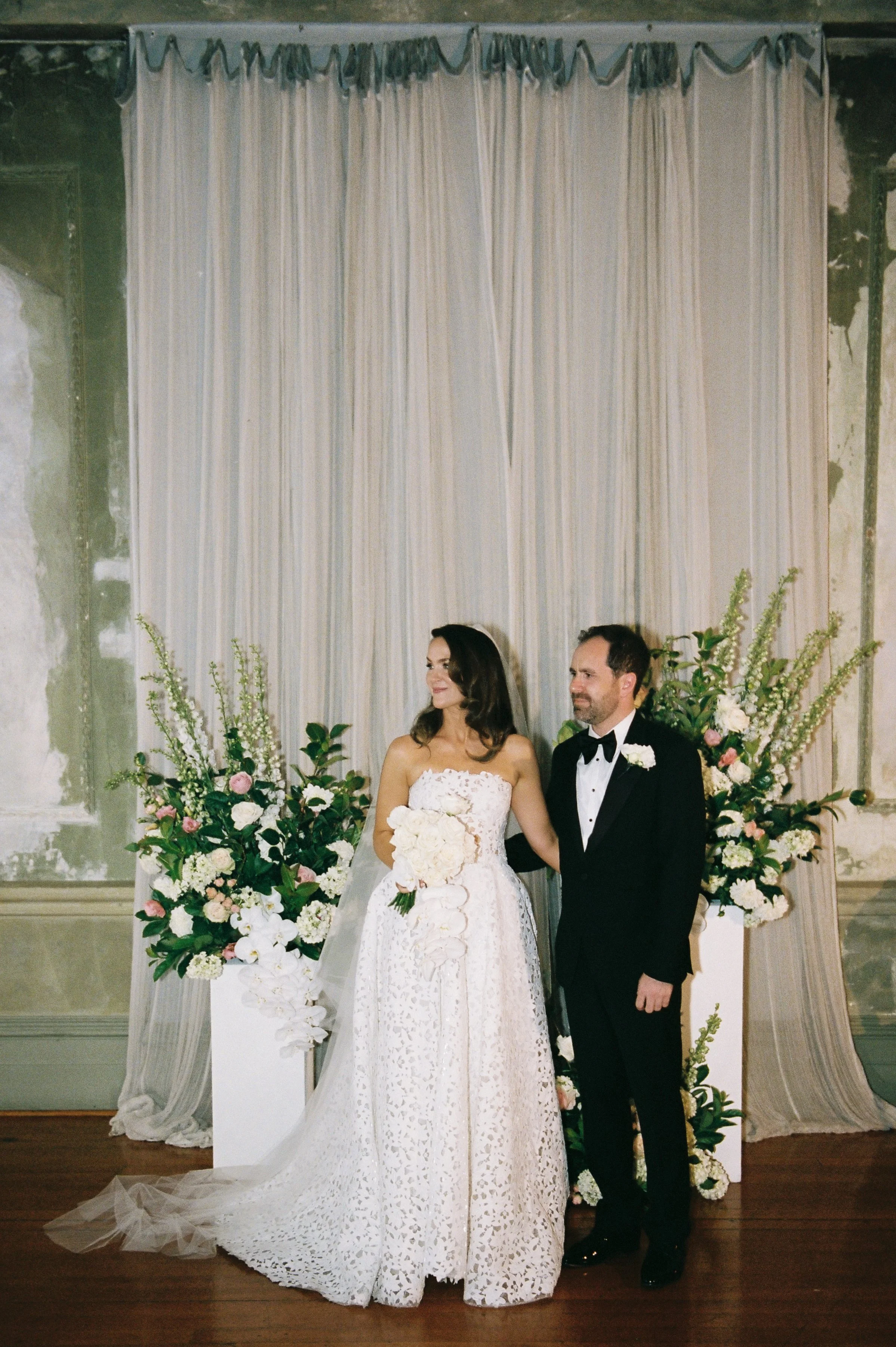 A bride and groom standing together at their wedding, surrounded by large floral arrangements with white, pink, and purple flowers, against a curtain backdrop.