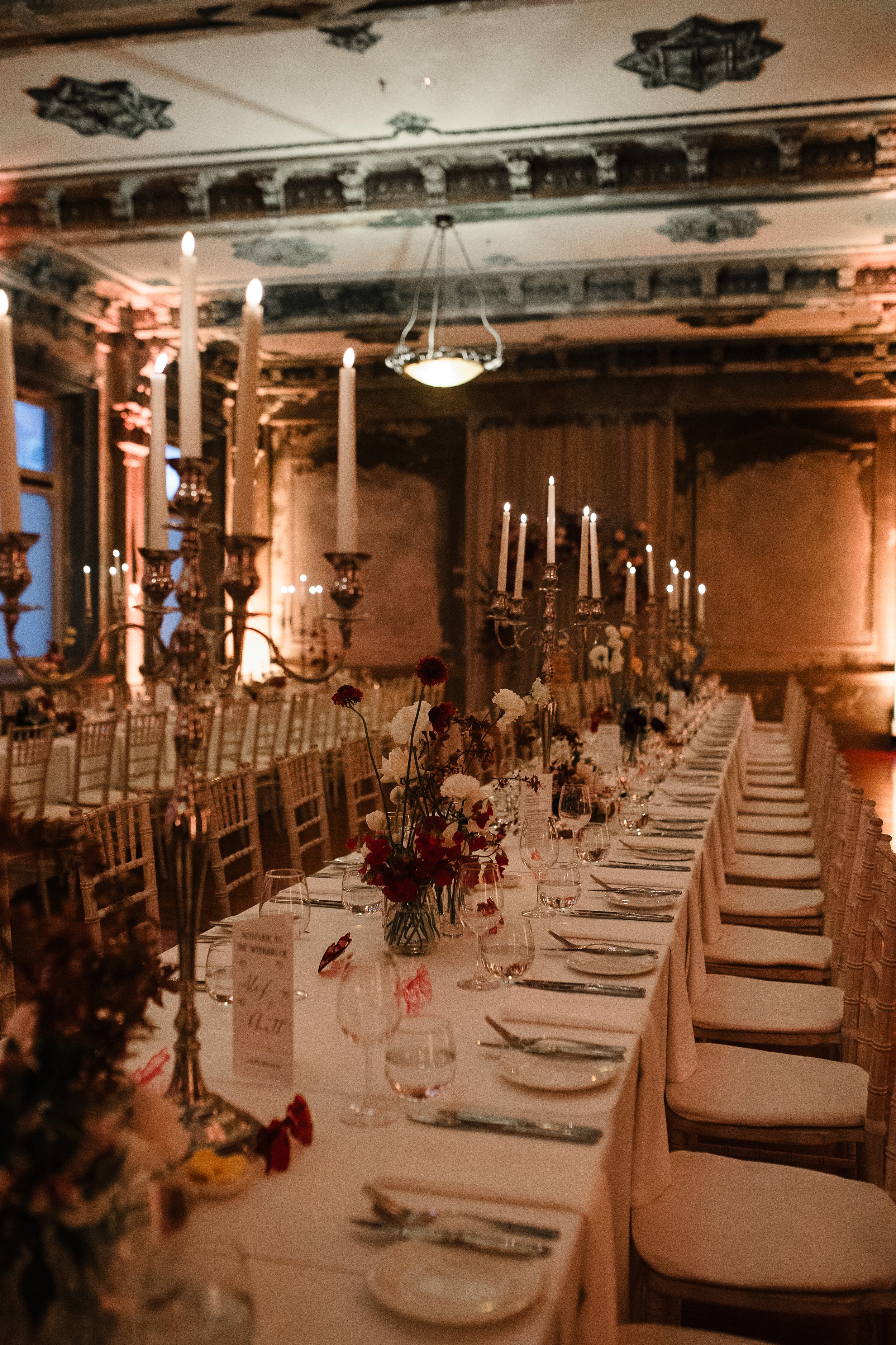 Long banquet table set with white tablecloth, floral centerpieces, and candles in a warmly lit elegant room with ornate ceiling details.