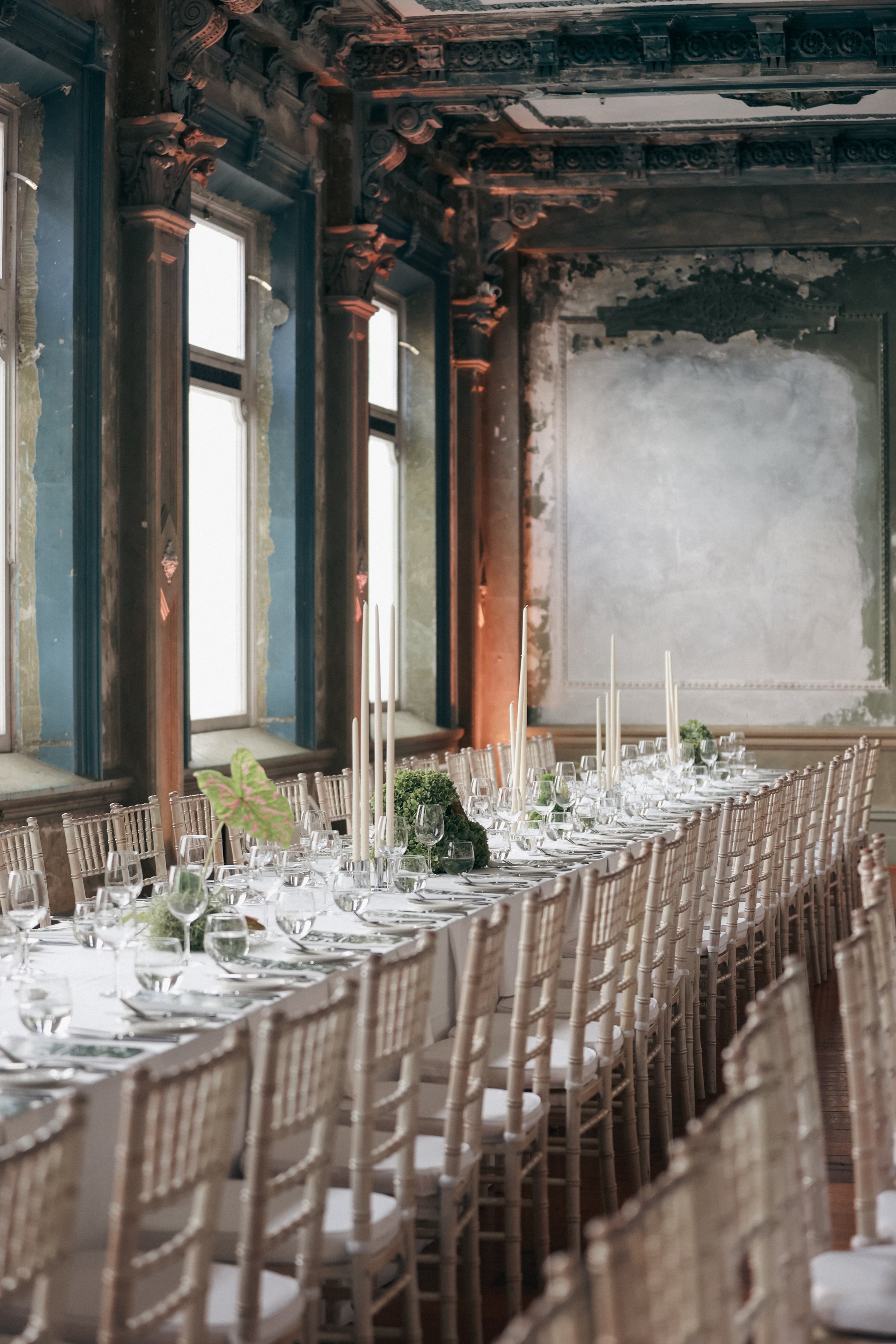 Long banquet table set with glassware, plates, and candles in an elegantly decorated room with tall windows and ornate architectural details.