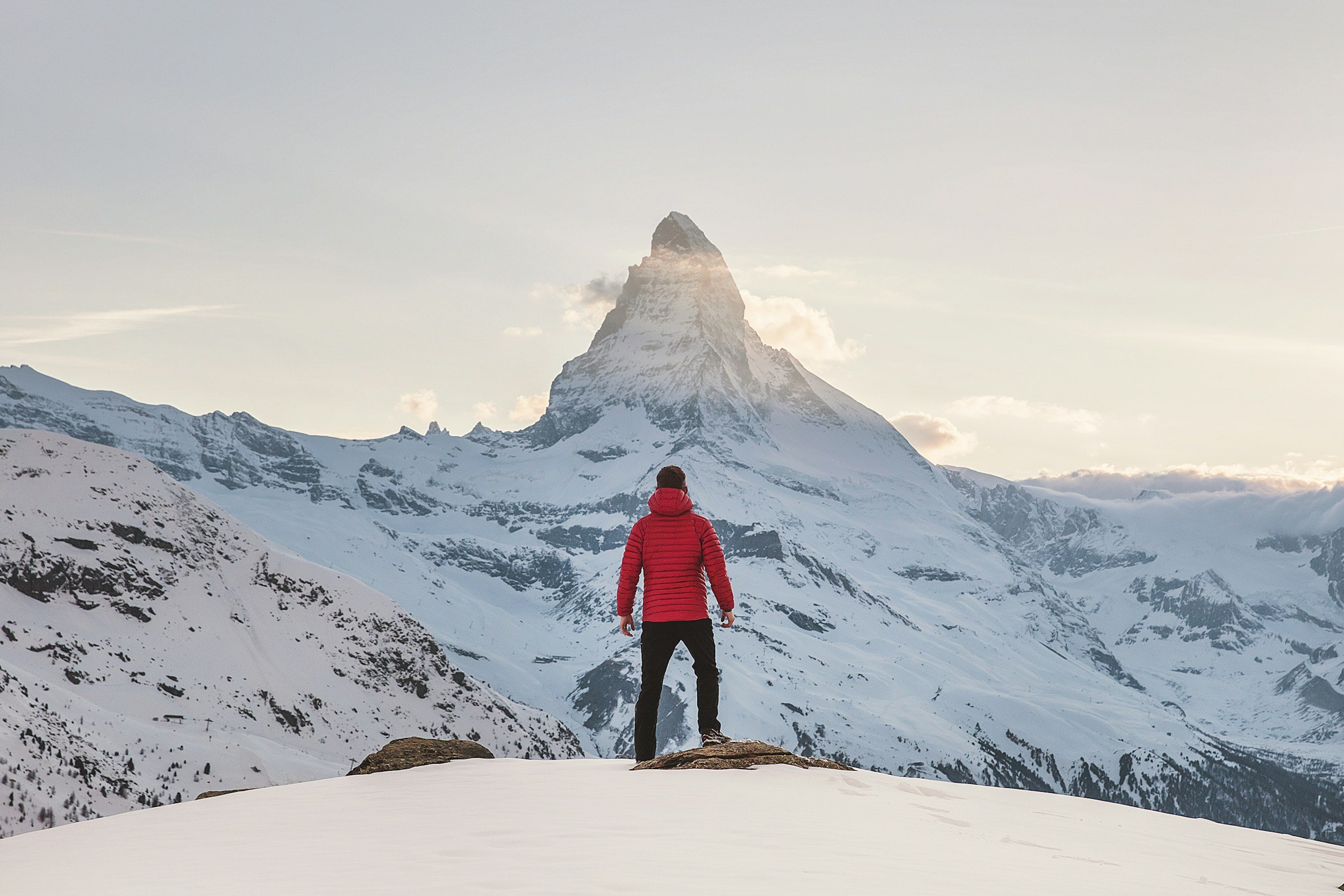 Person in red jacket standing on snowy ground, facing a snow-capped mountain with a pointed peak during daylight.