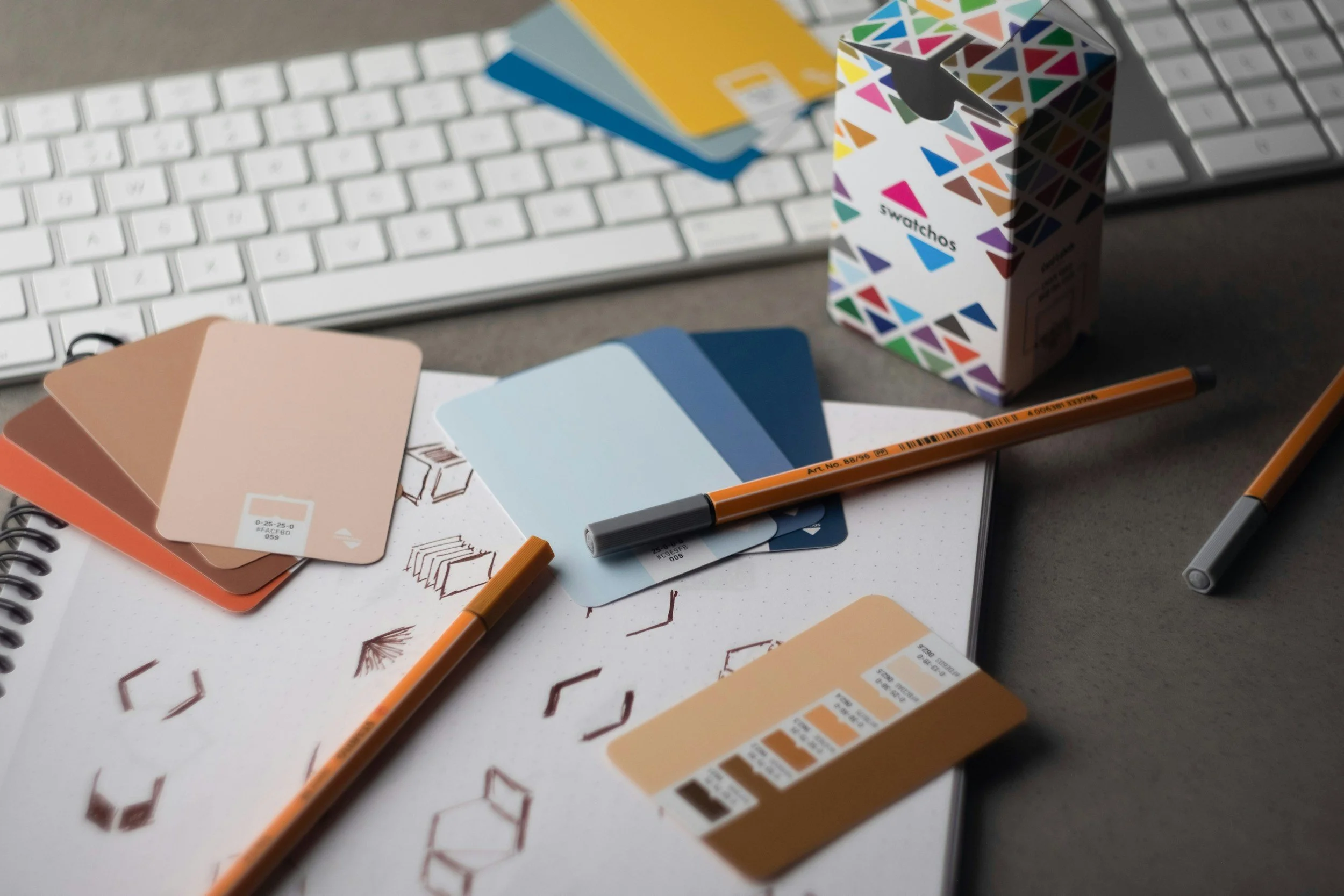 A workspace with color swatch cards, pens, a spiral notebook, and a keyboard on a desk.