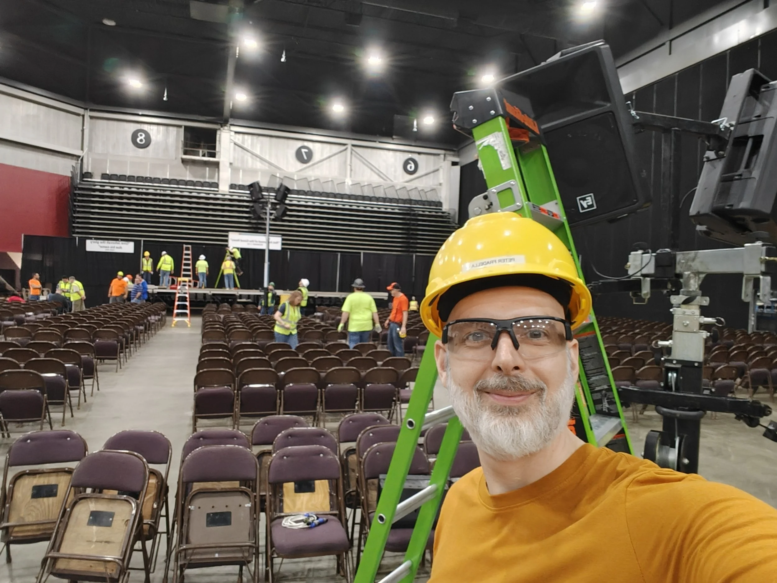 A man with gray hair and beard wearing safety glasses, a yellow hard hat, and an orange shirt taking a selfie inside an auditorium or event space. In the background, there are people working on setup, science seats, and equipment, including a large green and black lift.
