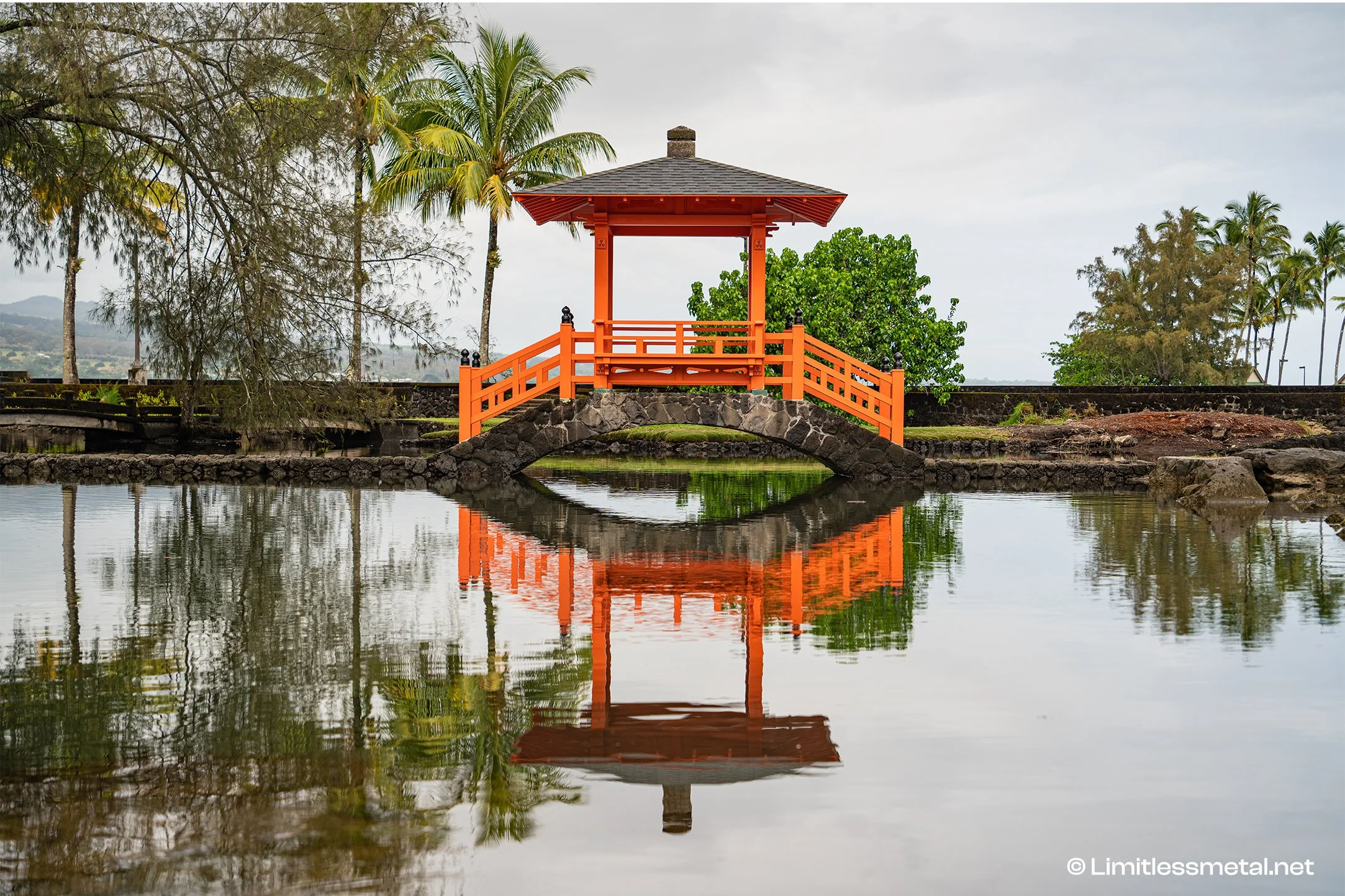 Serene fine art photograph of the iconic red Japanese bridge reflected in calm water at Liliʻuokalani Gardens in Hilo, Hawaii. Surrounded by tropical palms and lush greenery, this peaceful island landscape print brings balance, tranquility, and timel