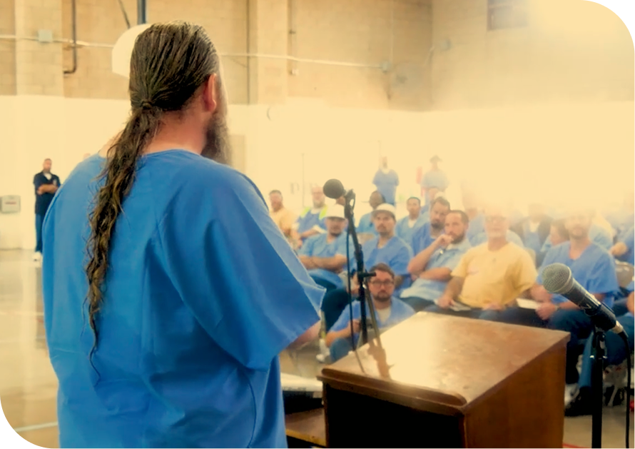 A person with long hair and a beard, dressed in a blue uniform speaking at a podium with microphones in a large indoor space, with an audience of people wearing similar blue uniforms and some in yellow, sitting and listening.
