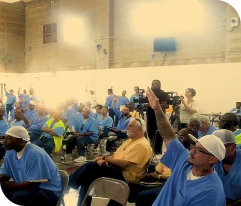 A large group of people seated indoors, some in blue uniforms, with one person raising their hand. A camera operator filming the event.