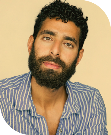 Close-up portrait of a man with curly dark hair, a full beard, and a mustache, wearing a blue and white striped shirt, looking at the camera against a neutral background.