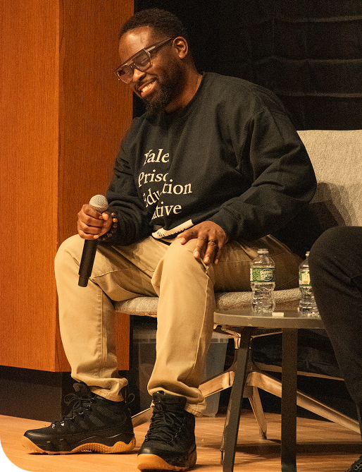 A man with glasses, a beard, and a black sweatshirt sitting on a chair, holding a microphone, smiling, with water bottles on a small table beside him.