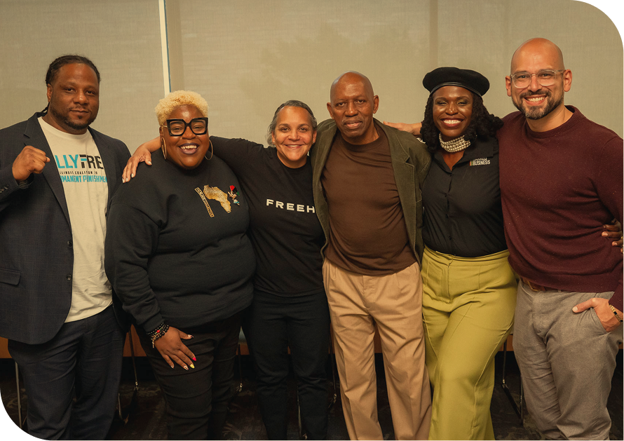 Group of six diverse people standing together, smiling, with arms around each other, in an indoor setting.