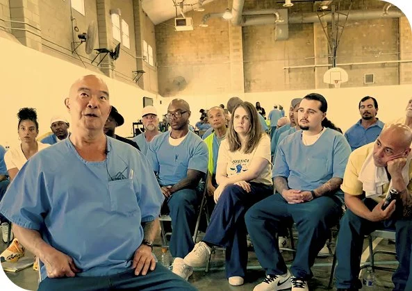 A diverse group of people sitting on chairs in a gymnasium or large indoor space, attentively listening to a speaker.