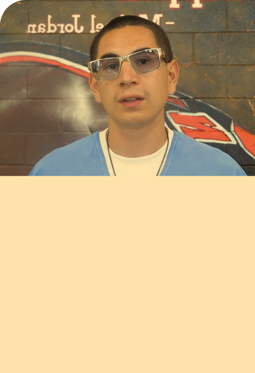 A young man with short dark hair, wearing glasses, a white T-shirt, a light blue zip-up jacket, and a necklace, stands in front of a brick wall with partially visible red and black text or logo.