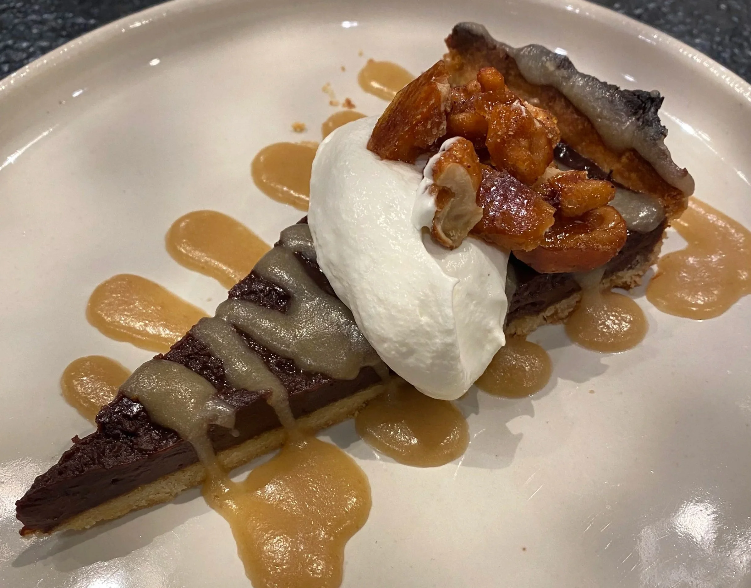 Slice of chocolate tart topped with whipped cream, caramelized nuts, and caramel sauce on a white plate.