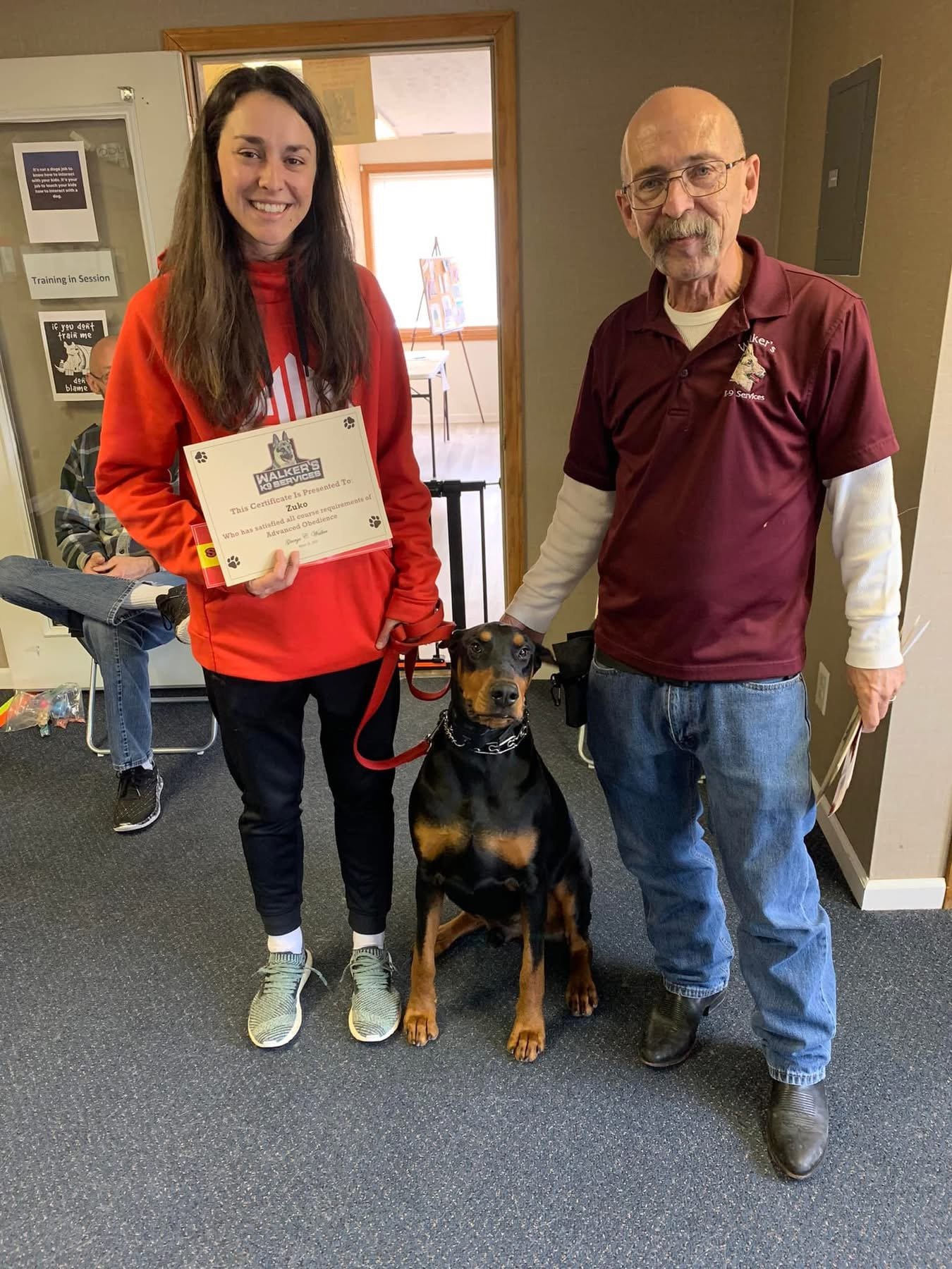 A young woman holding a certificate, a man, and a black and tan dog sitting between them indoors. The woman is wearing a red hoodie and the man is wearing a maroon shirt with a logo. In the background, a person is seated and there are posters on the wall.