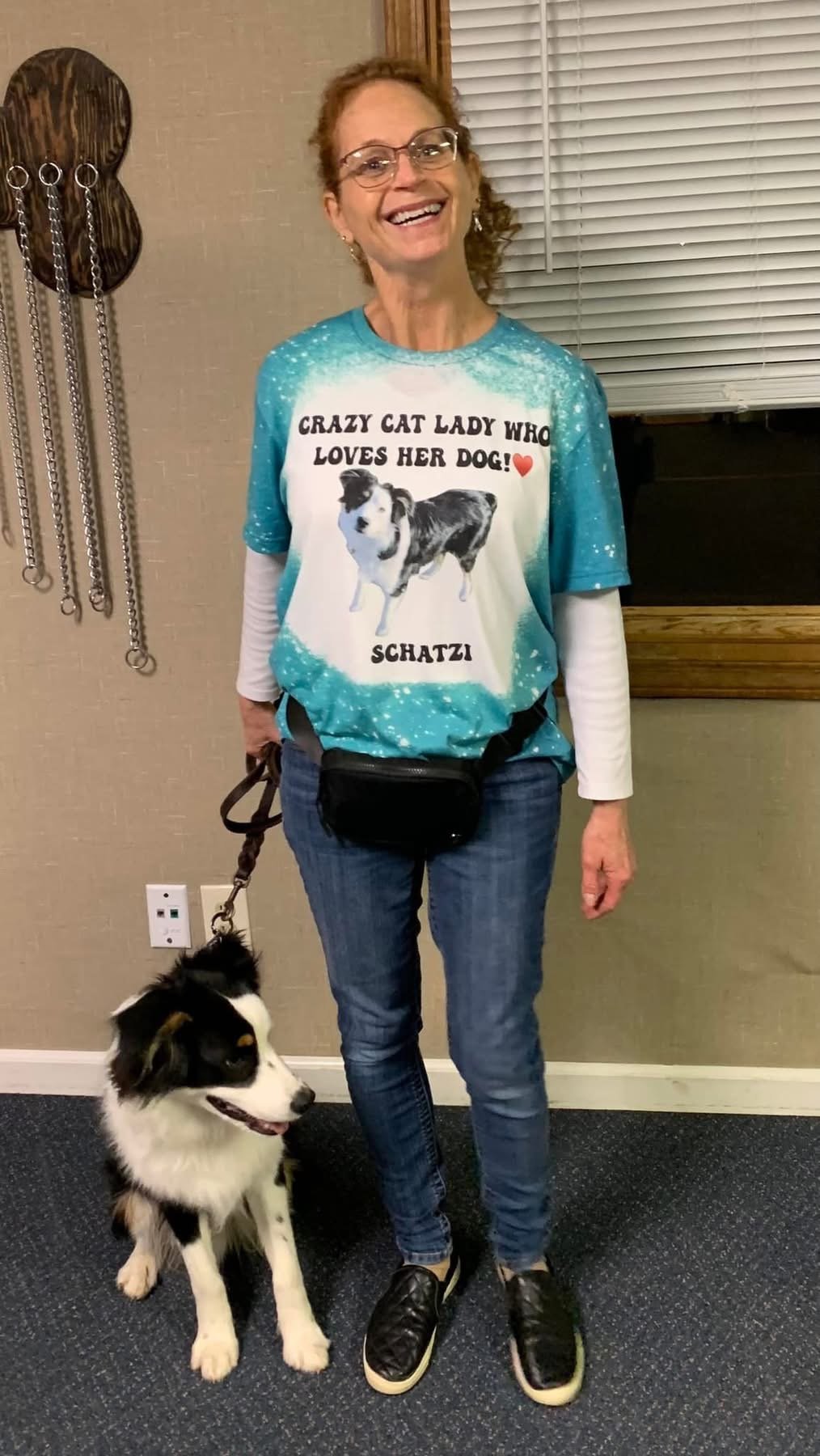 A woman with glasses and curly red hair standing indoors, smiling, holding a leash with a black and white Border Collie. She is wearing a blue tie-dye T-shirt with a dog photo and text, and jeans.