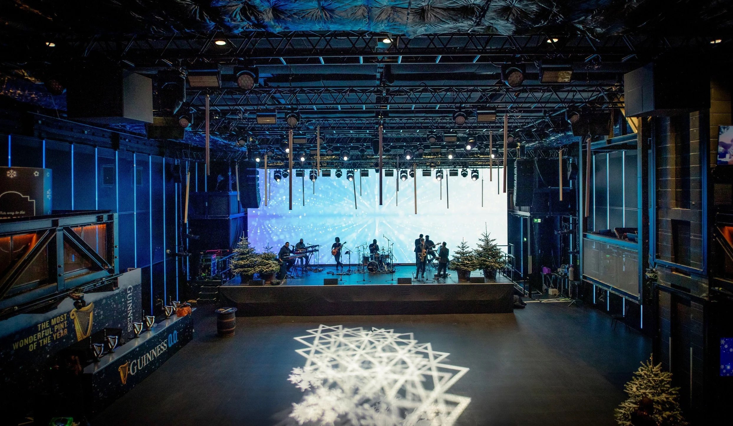 Indoor stage set up for a live band performance with Christmas trees and festive decorations, illuminated by a lights and snowflake projections.