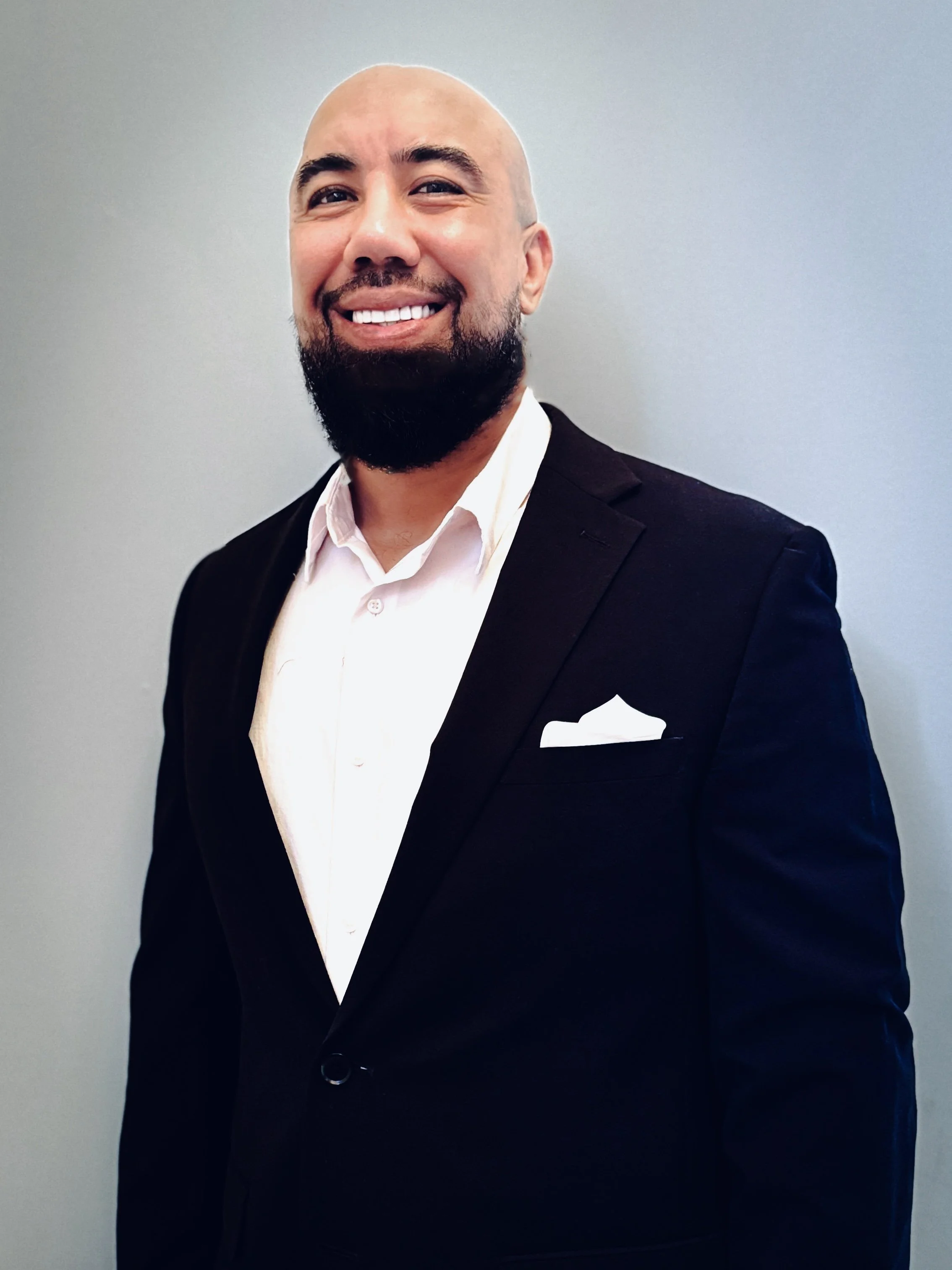 A smiling man with a beard and a shaved head wearing a black suit jacket over a white dress shirt, standing against a plain gray background.