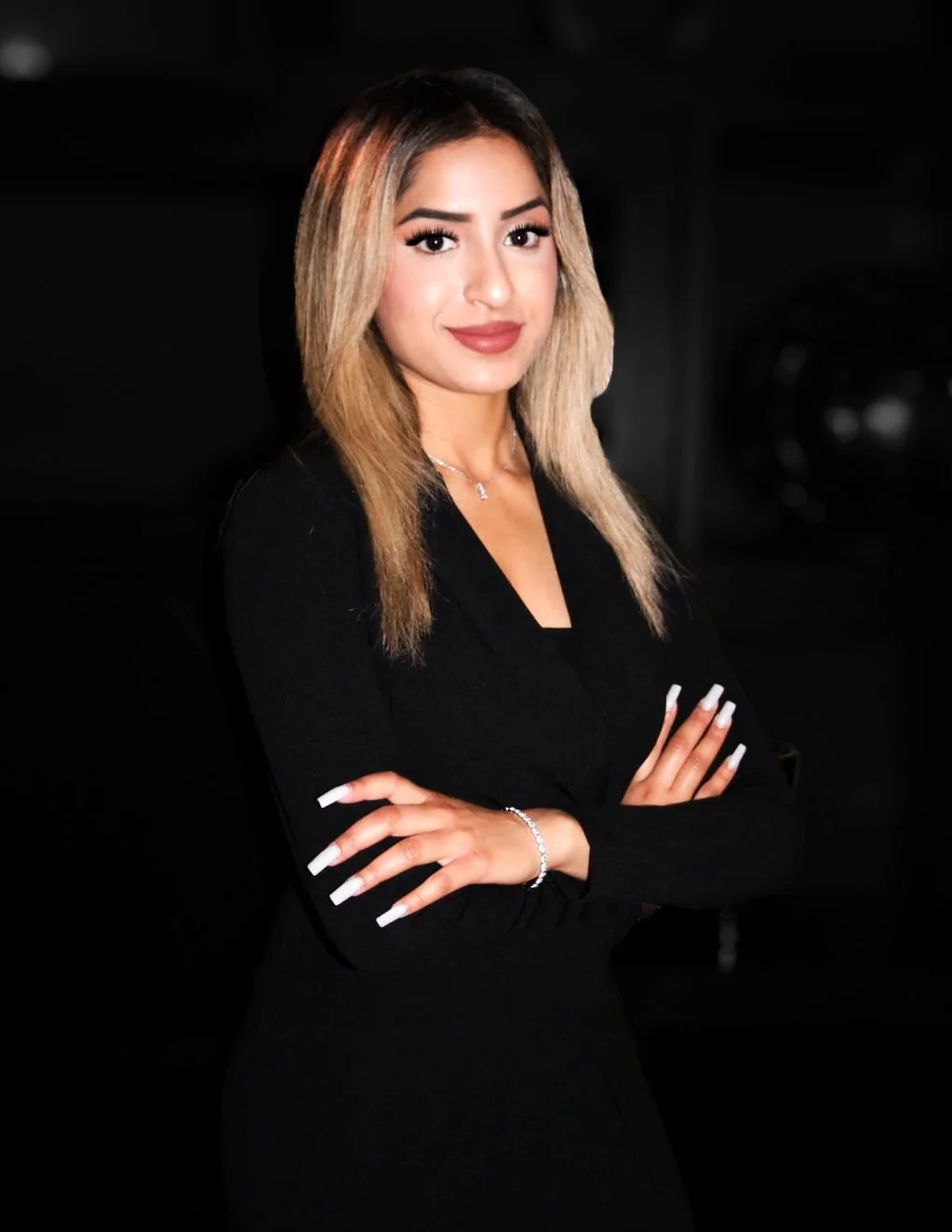 A woman with blonde hair, wearing a black top, and jewelry, standing with arms crossed against a black background.