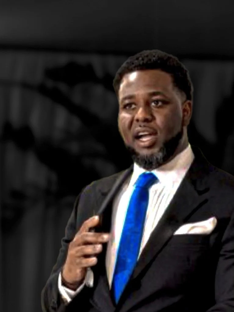 A man with dark skin, short black hair, and a beard, dressed in a dark suit with a white shirt and blue tie, speaking and holding a microphone against a blurry dark background.