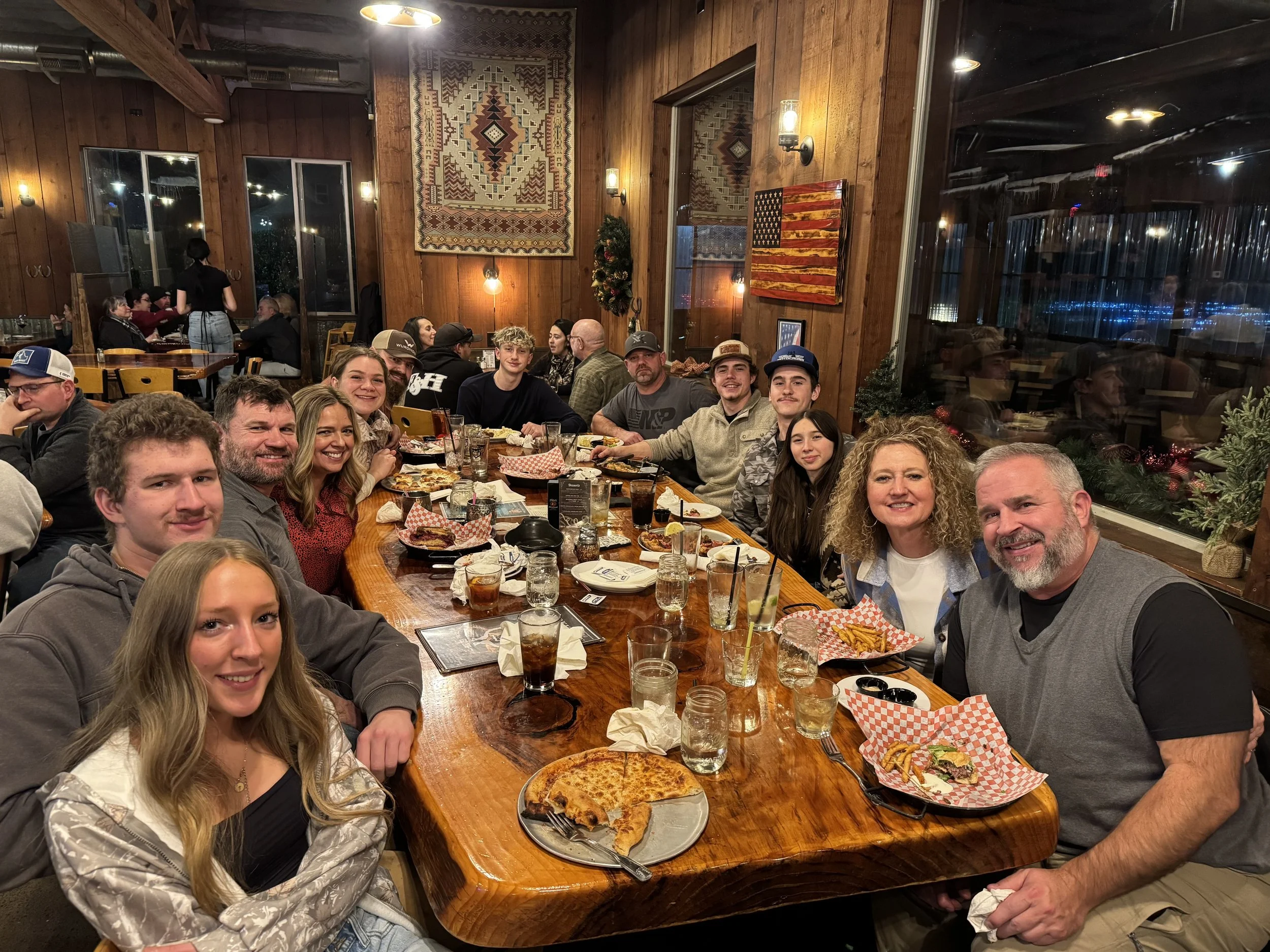 A large group of people sitting around a long wooden table in a restaurant, enjoying food and drinks, with some people smiling and others engaged in conversations. The restaurant has wood-paneled walls, decorative wall art, and warm lighting.