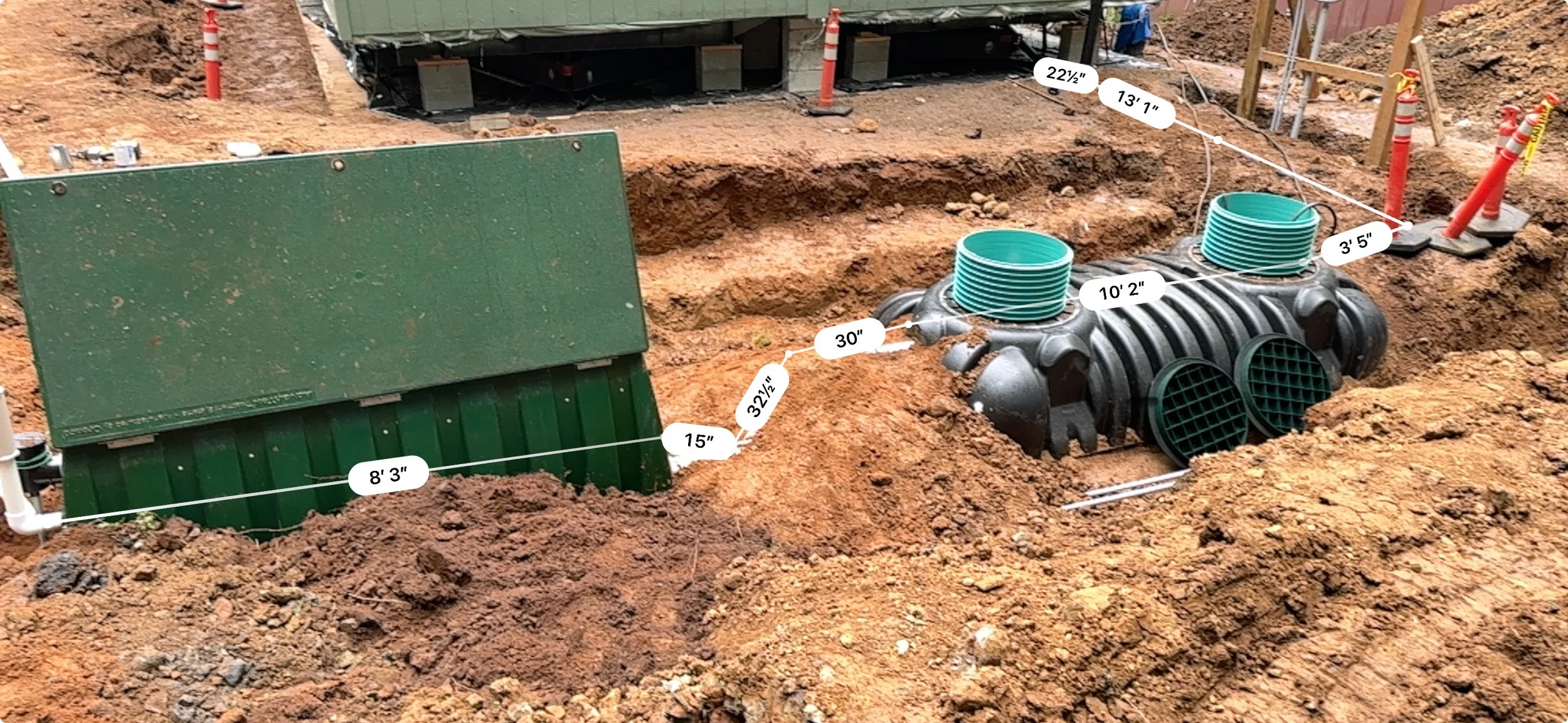 Construction site with underground drainage system, showing measurement labels for pipes and fittings in a trench with bare soil, orange cones, and construction materials.