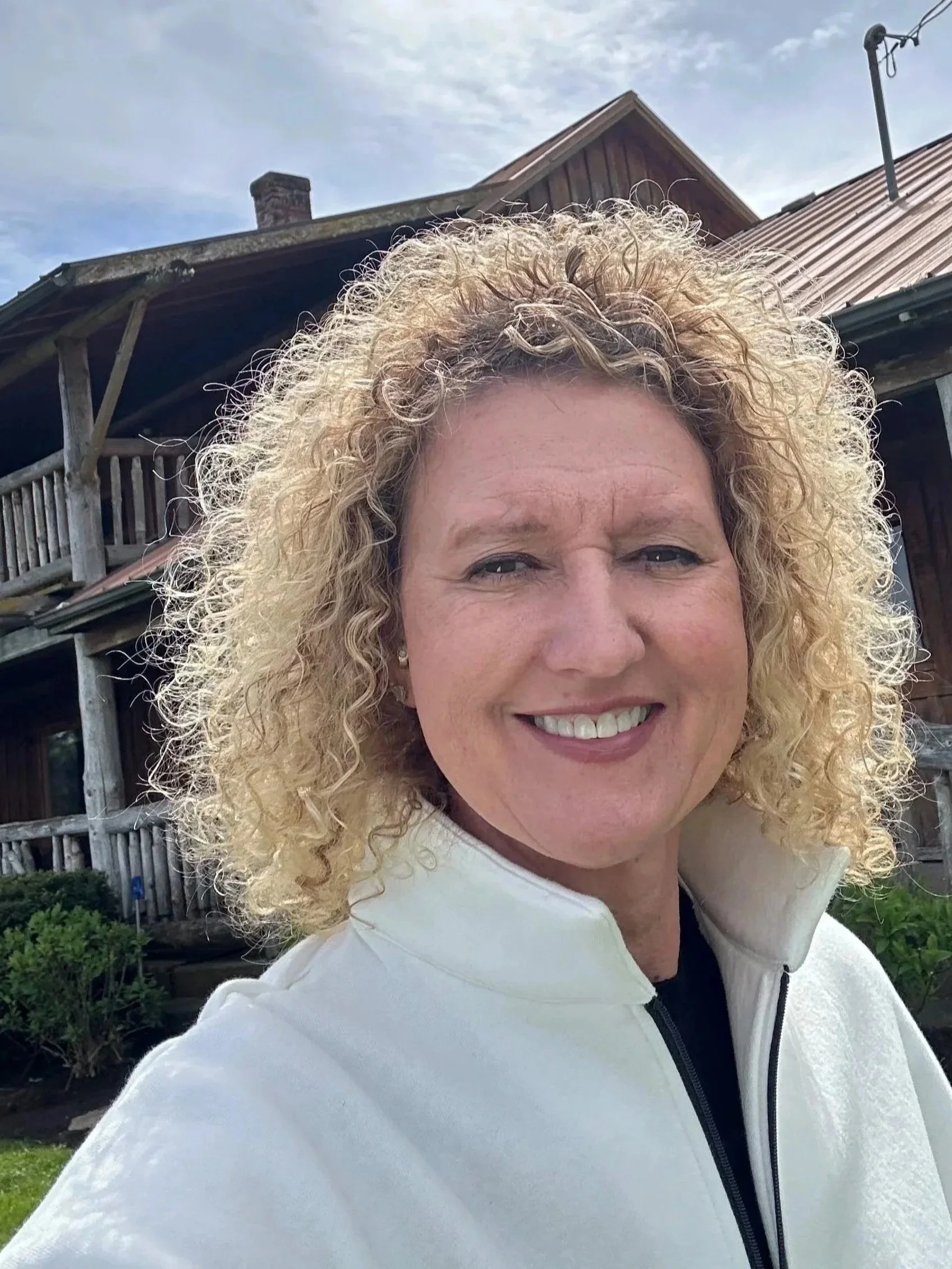 A woman with curly blonde hair smiling outdoors in front of a rustic wooden house with a balcony and a chimney.