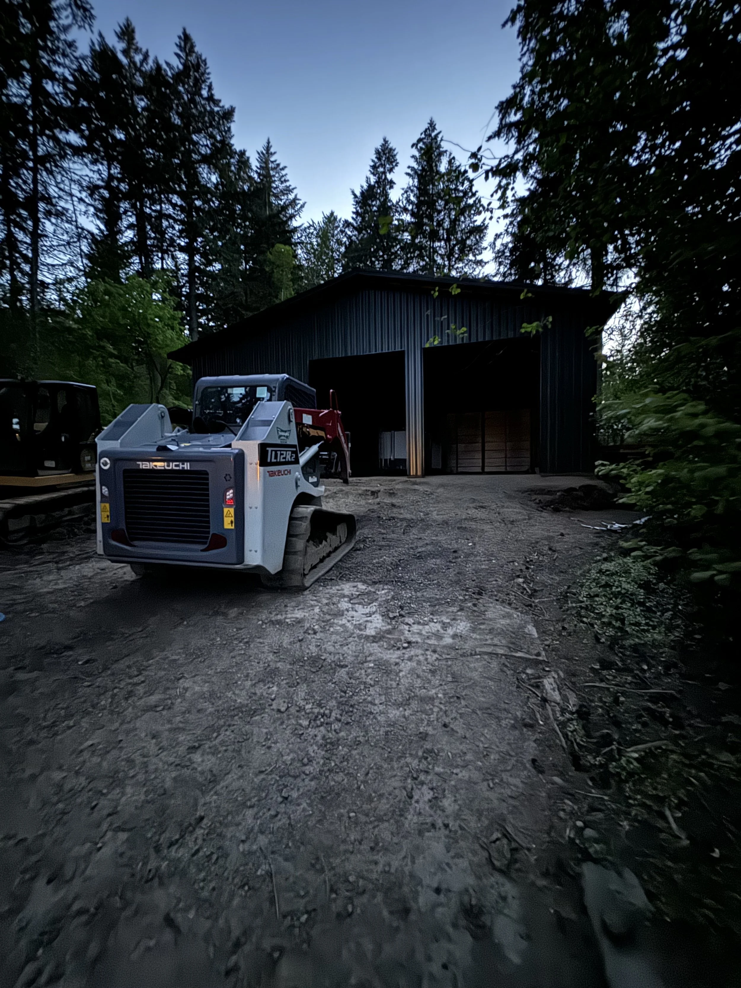 A small construction vehicle parked on a dirt path outside a dark metal barn surrounded by trees during twilight.