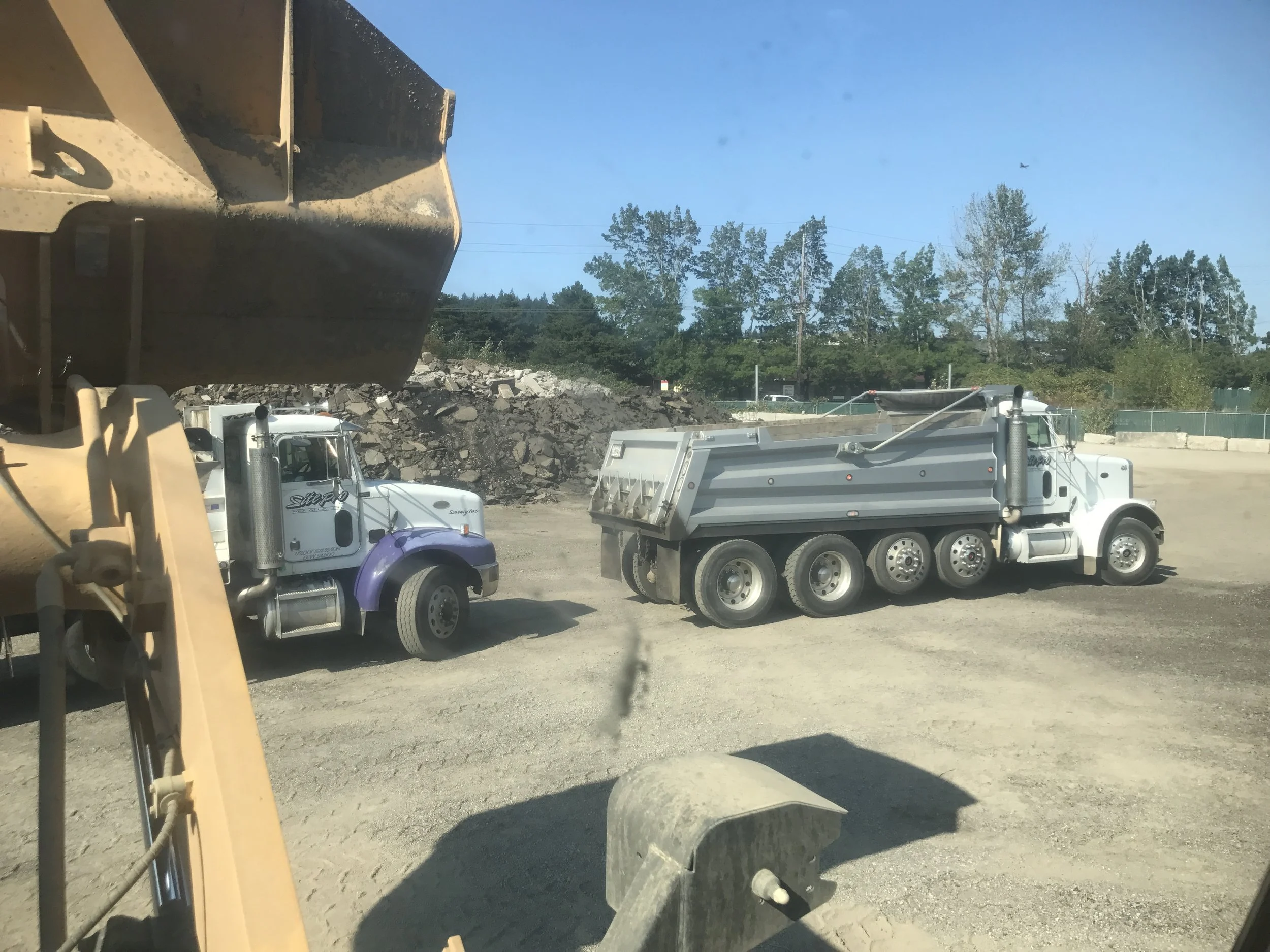 Two dump trucks on a construction site, one partially visible in the foreground and the other in the background, with gravel and dirt piles behind them and trees in the distance.