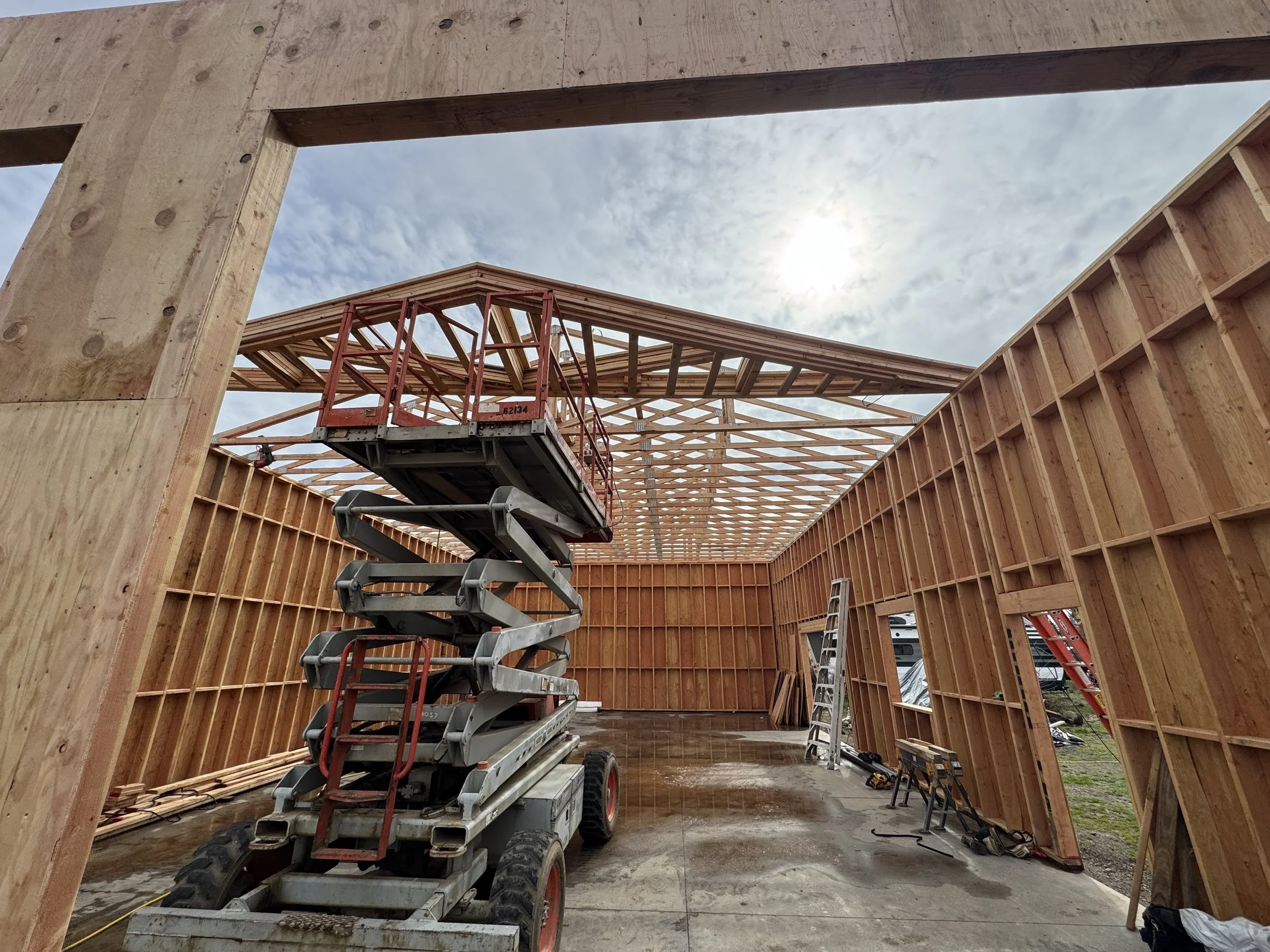 Construction site in a building framework with wooden walls and roof trusses, a scissor lift, ladder, tools, and a cloudy sky.