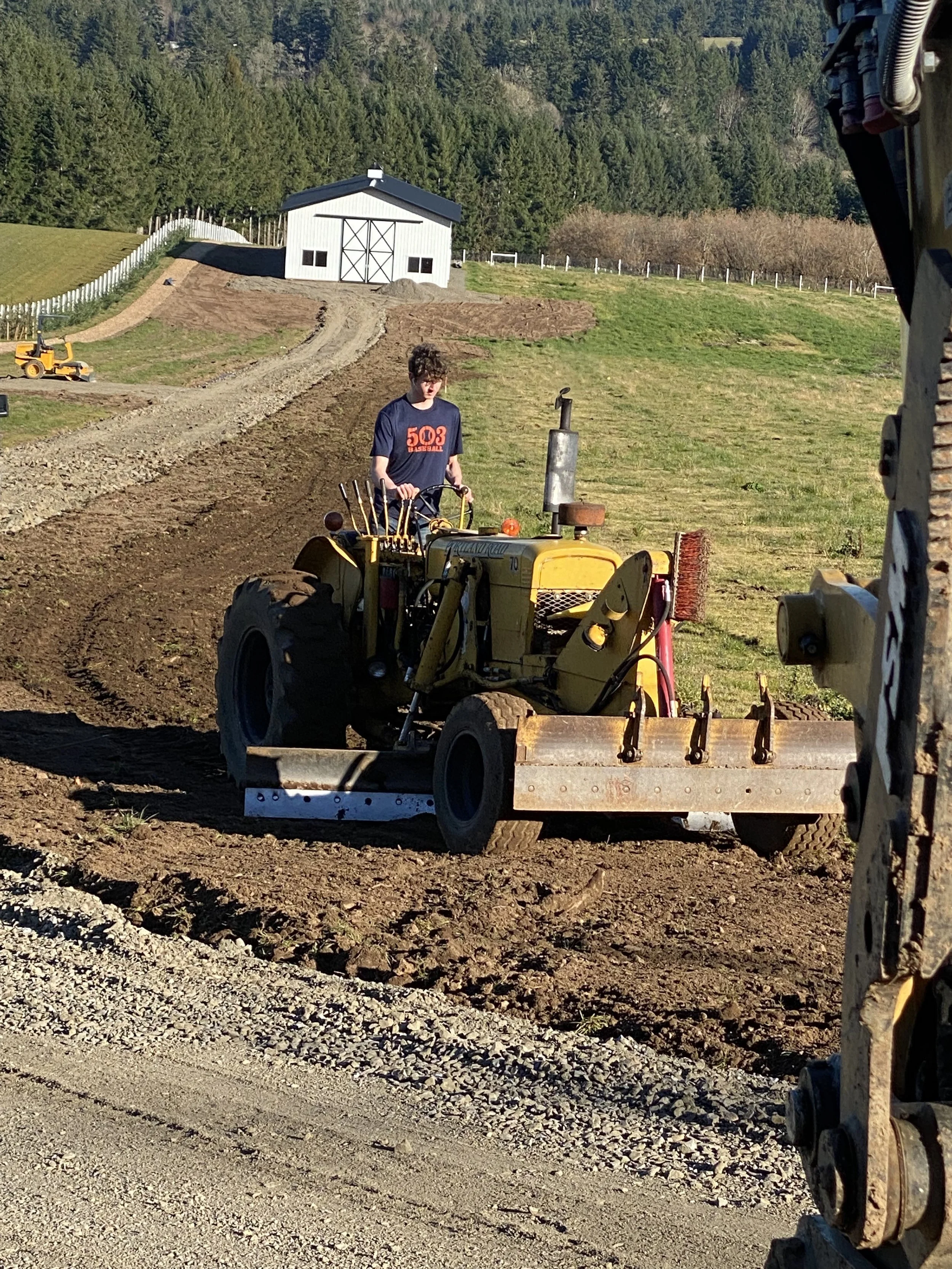 A person operating a yellow tractor on a construction site with a sloped dirt pathway, green grass, a white building in the background, and surrounding trees.