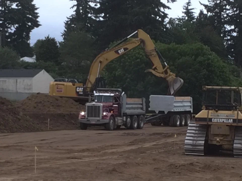 Construction site with a large yellow excavator loading dirt into a red dump truck, and a yellow bulldozer nearby, with trees and a small building in the background.