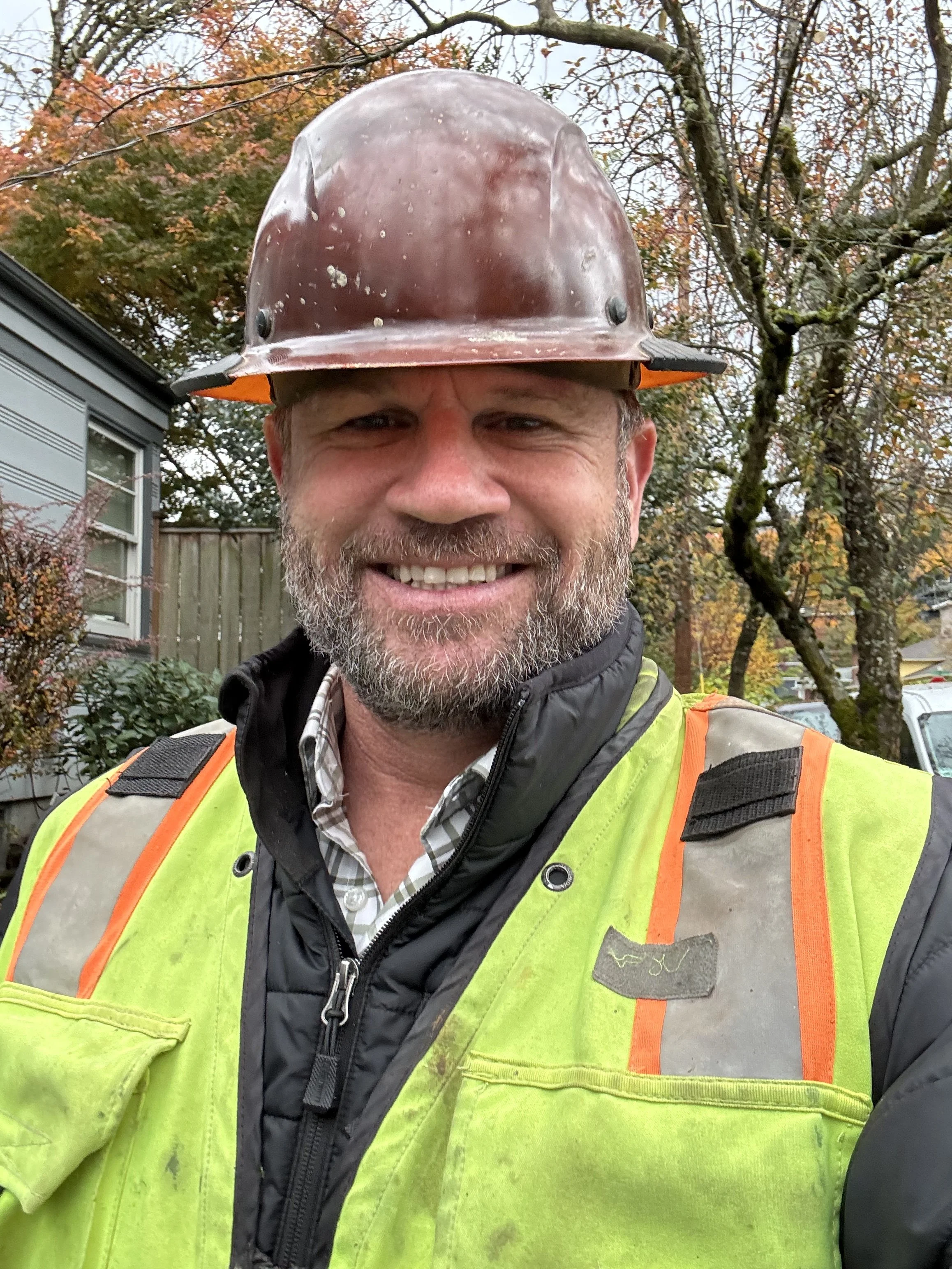 A man smiling outdoors, wearing a brown hard hat and a neon green safety vest over a black jacket and plaid shirt, with trees and a house in the background.