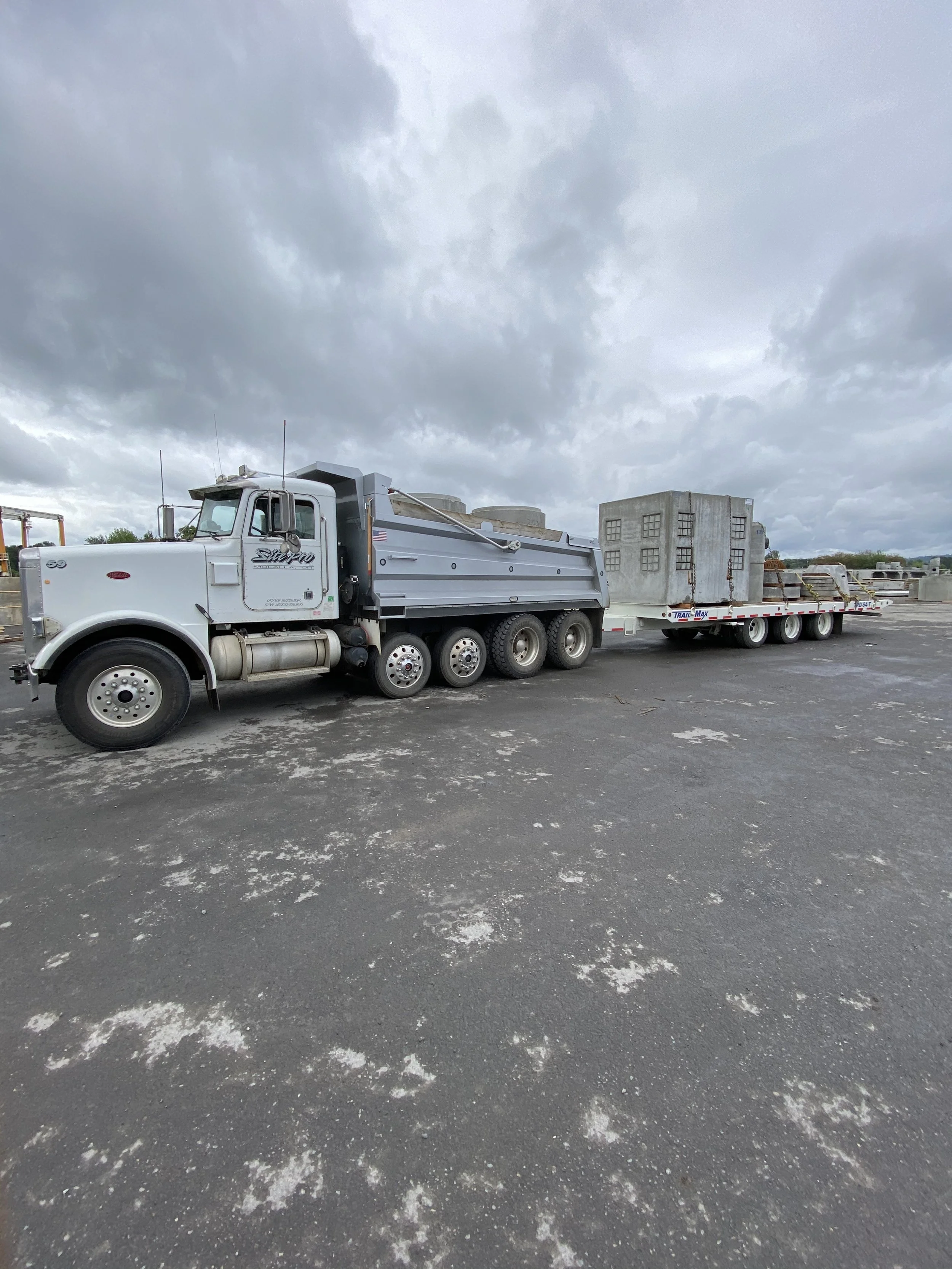 A large white truck with a grey trailer parked on a paved lot under a cloudy sky, with other trailers and equipment in the background.