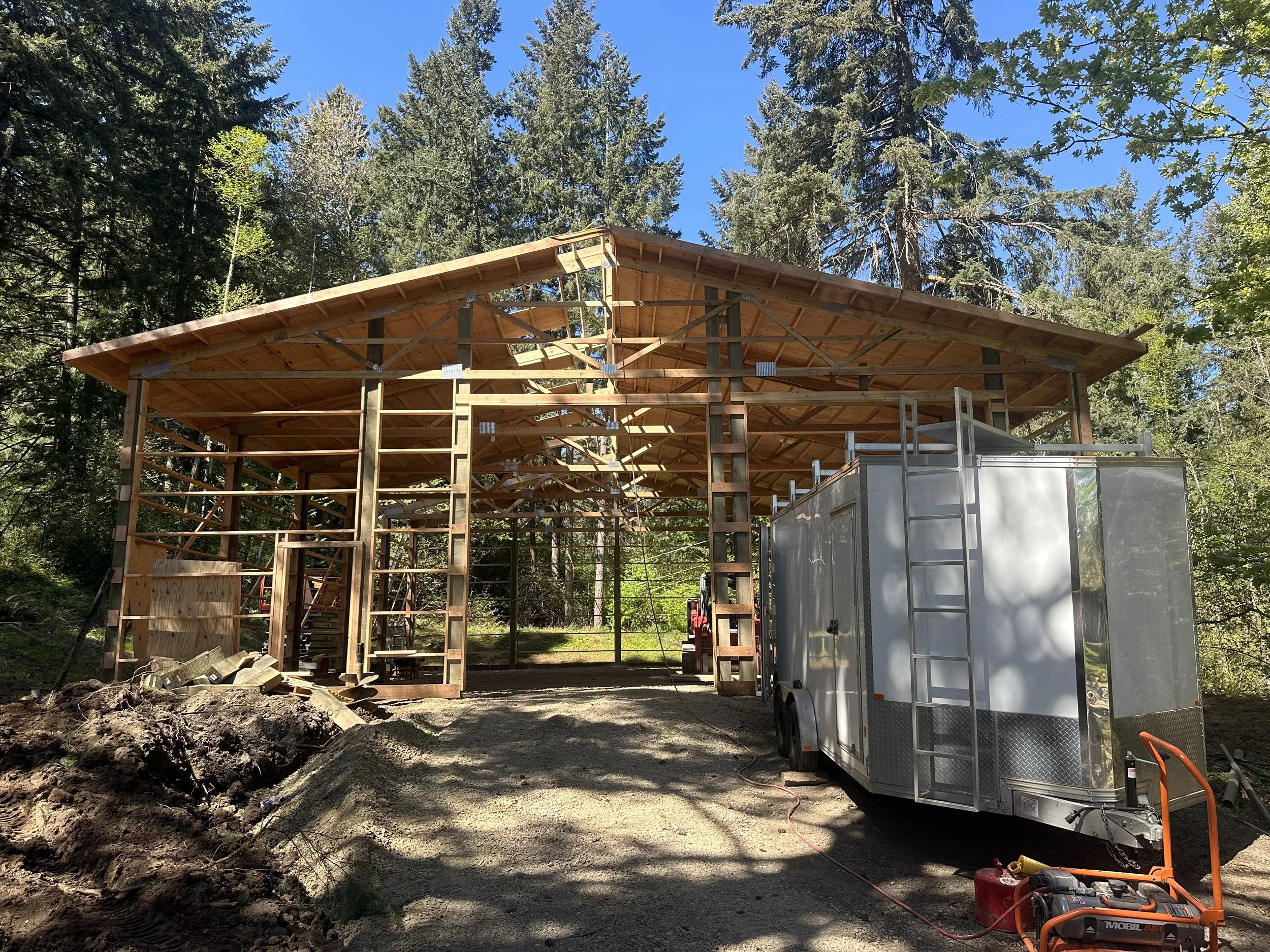 Under construction wooden building in a forested area with scaffolding, a trailer, and construction tools.